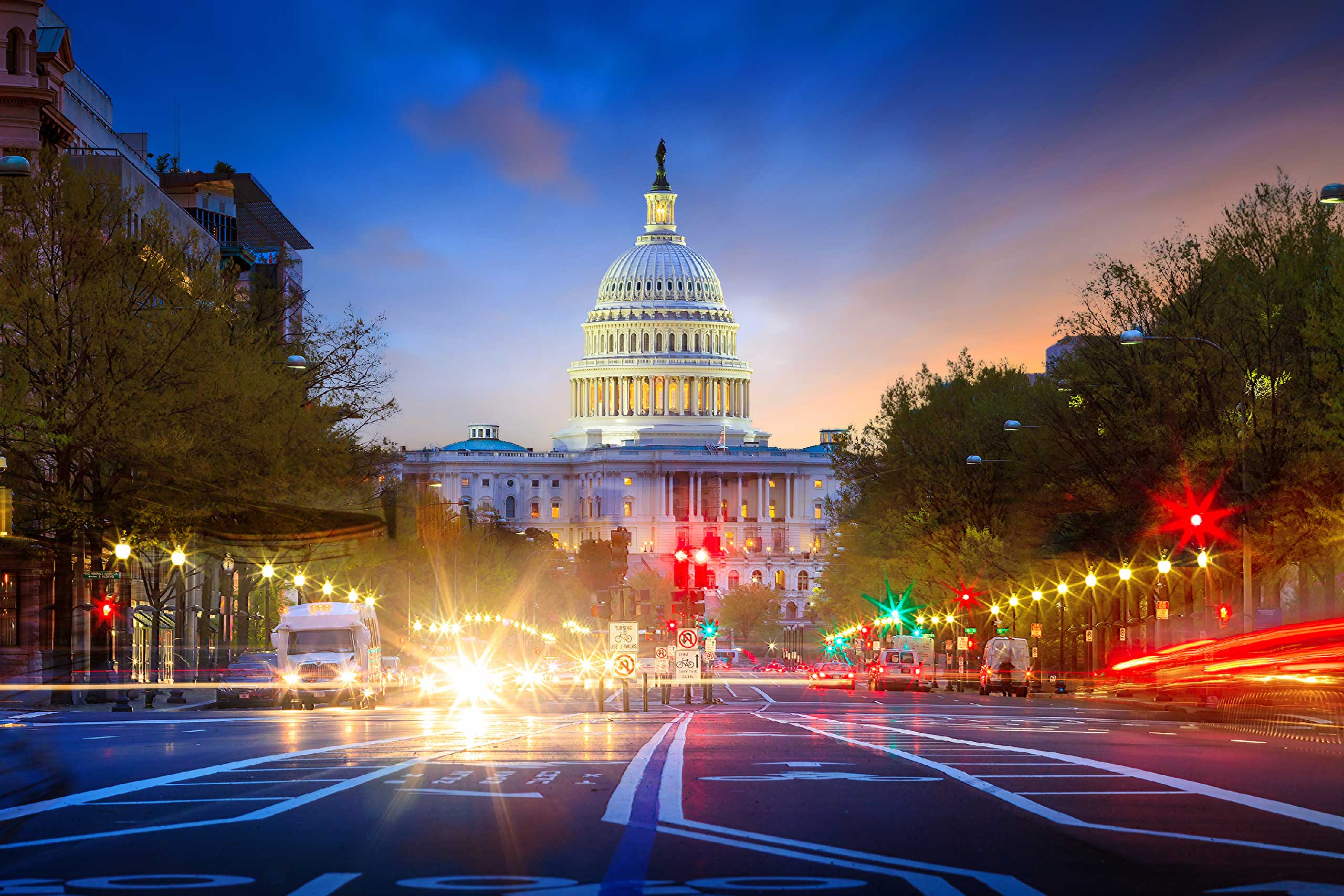 The U.S. Capitol illuminated at dusk, framed by vibrant city lights and bustling street activity.