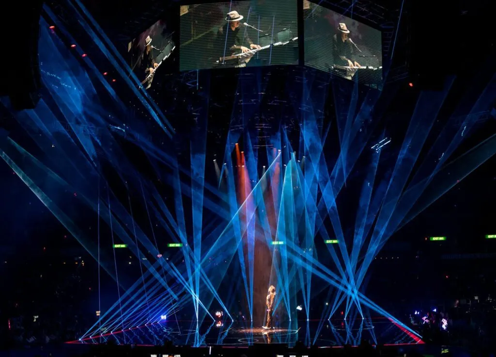 A musician performs on a stage surrounded by vibrant blue laser lights and large screen displays.