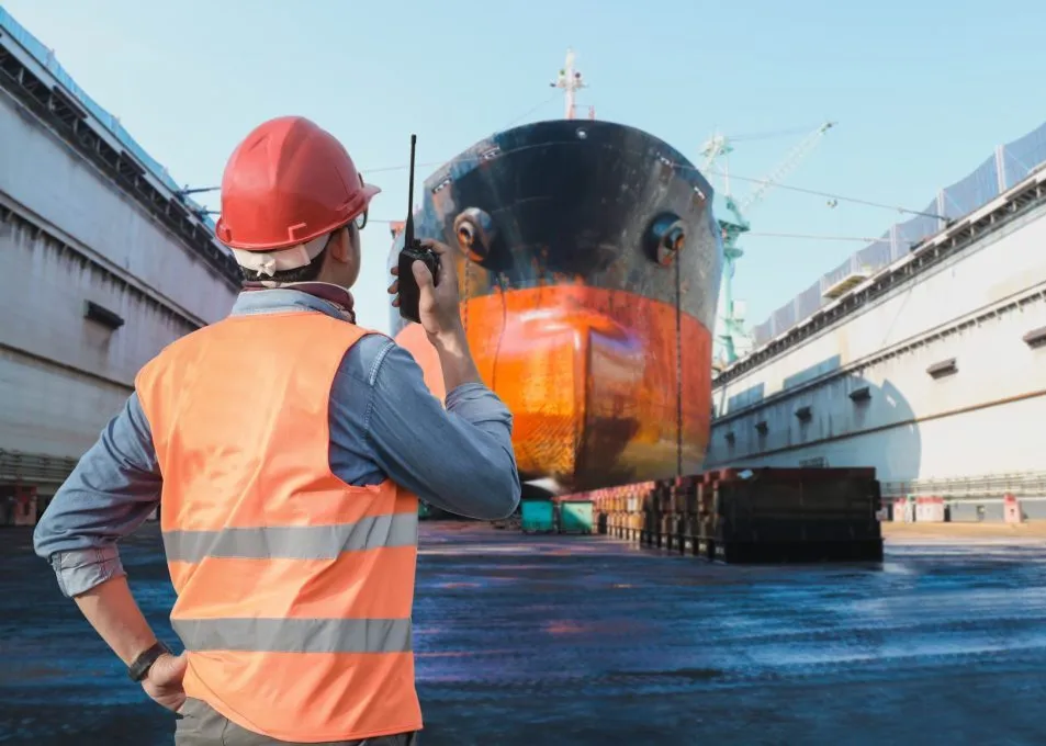 A worker in safety gear oversees a large ship in a dry dock at a shipyard.