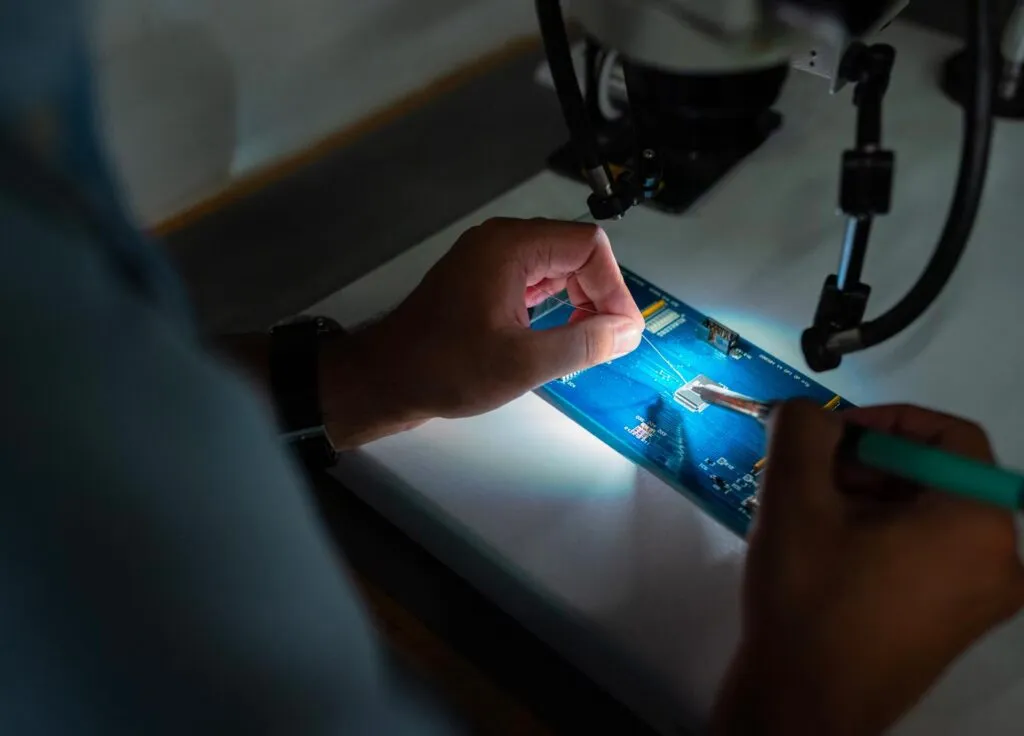 A person works on a circuit board under a microscope with precision tools and focused lighting.