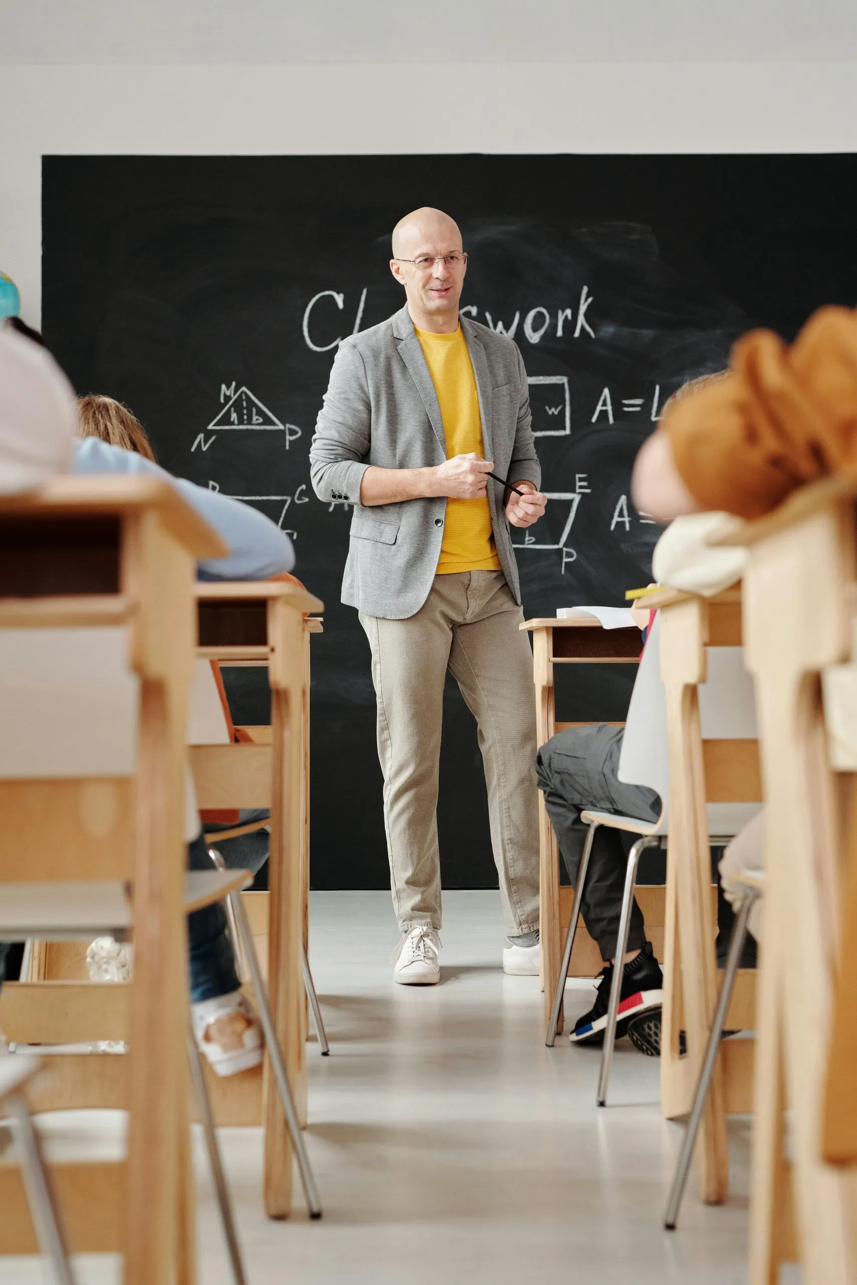 A teacher stands in a classroom near a blackboard with math equations, addressing seated students.