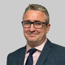 A man in a suit and tie, wearing glasses, stands against a plain gray background.