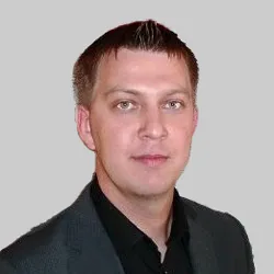A man wearing a dark suit jacket and shirt stands against a plain gray background.