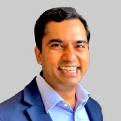 A man in a formal suit and light blue shirt is posed against a plain gray background.