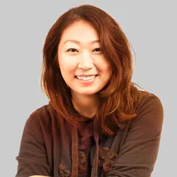 A person with shoulder-length brown hair wearing a dark hoodie against a gray background.