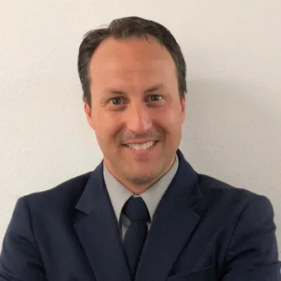 A person wearing a dark suit, light shirt, and tie stands against a plain white background.