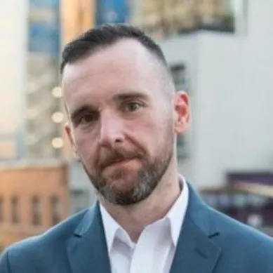 A person in a suit stands outdoors with an urban background featuring buildings and soft lighting.