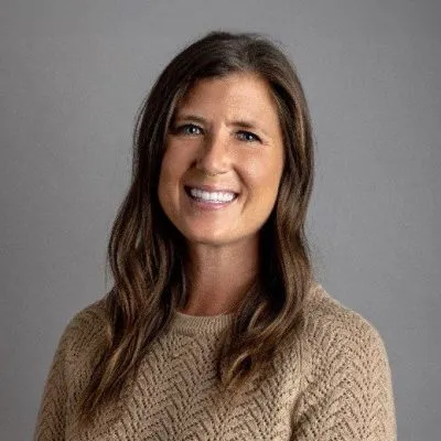 A woman with long, wavy brown hair wearing a textured beige sweater against a gray background.