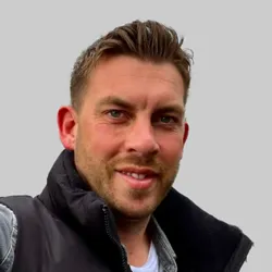 A man wearing a black vest over a gray shirt stands against a plain gray background.