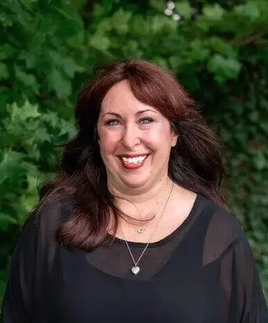 A woman with long, dark hair wears a black top and a necklace, standing outdoors near green foliage.