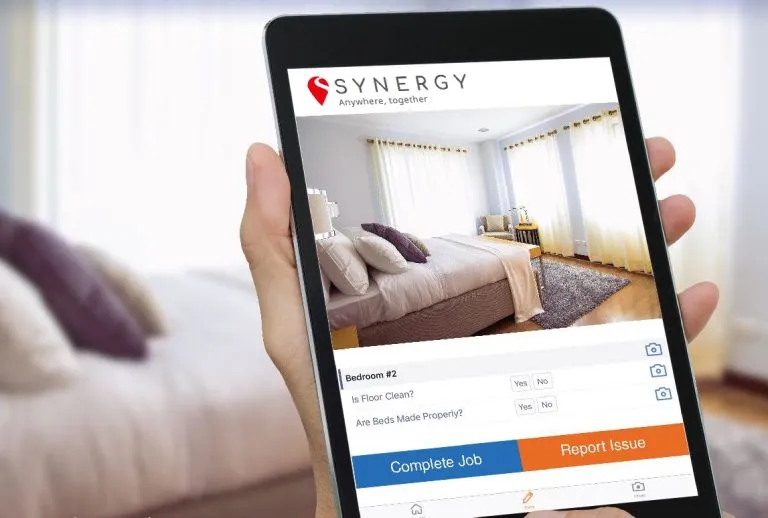 A tablet displays a cleaning checklist app by Synergy, with a bedroom photo in the background.