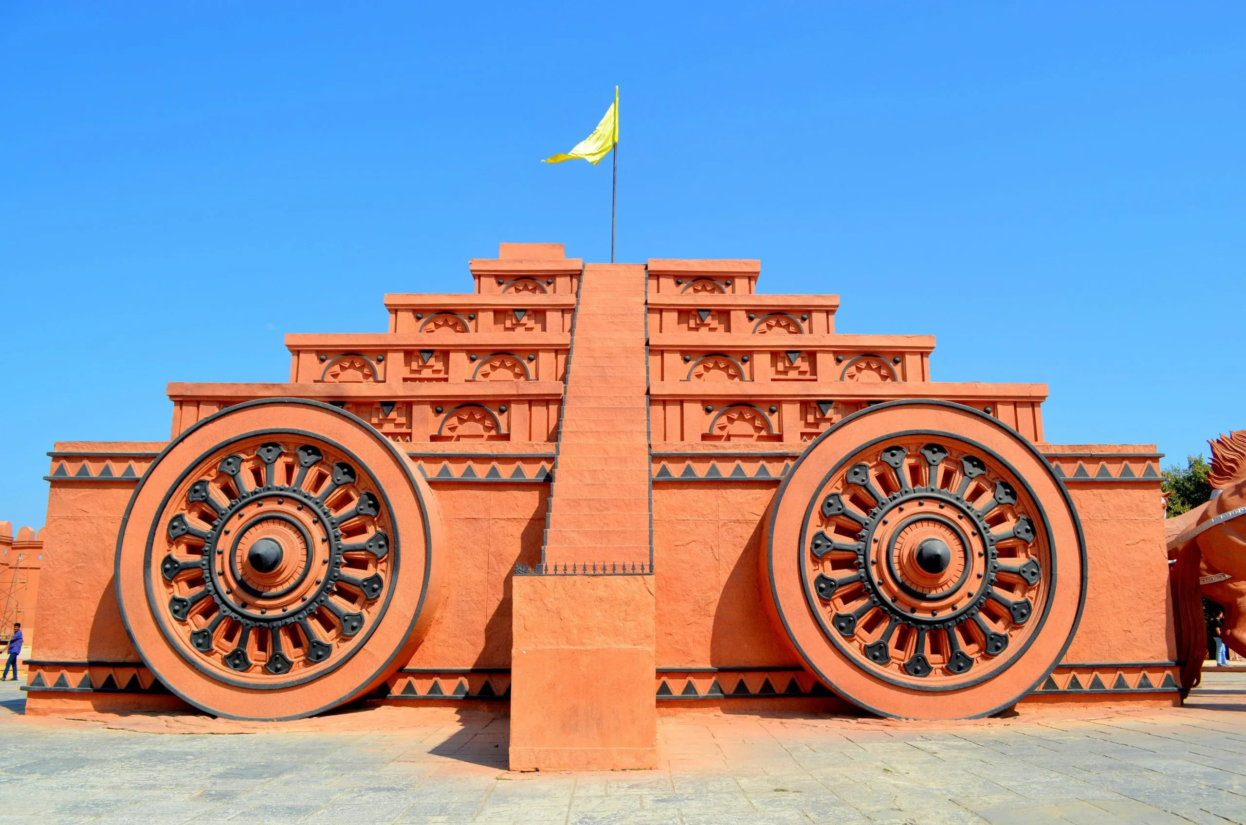 A terracotta chariot structure with intricate wheels and a yellow flag atop, under a clear blue sky.
