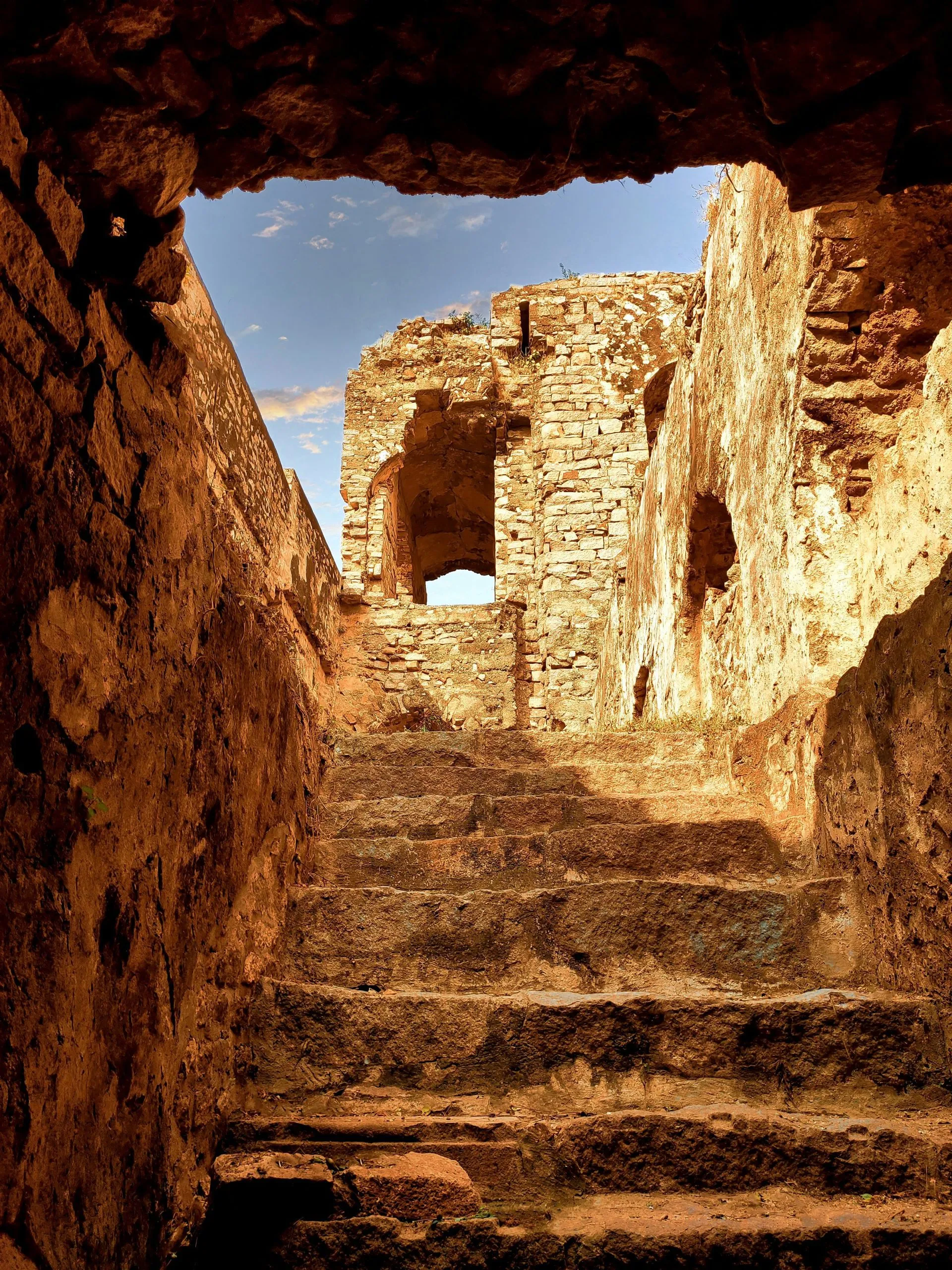 Stone steps lead to ancient ruins under a bright sky, framed by rugged walls and archways.