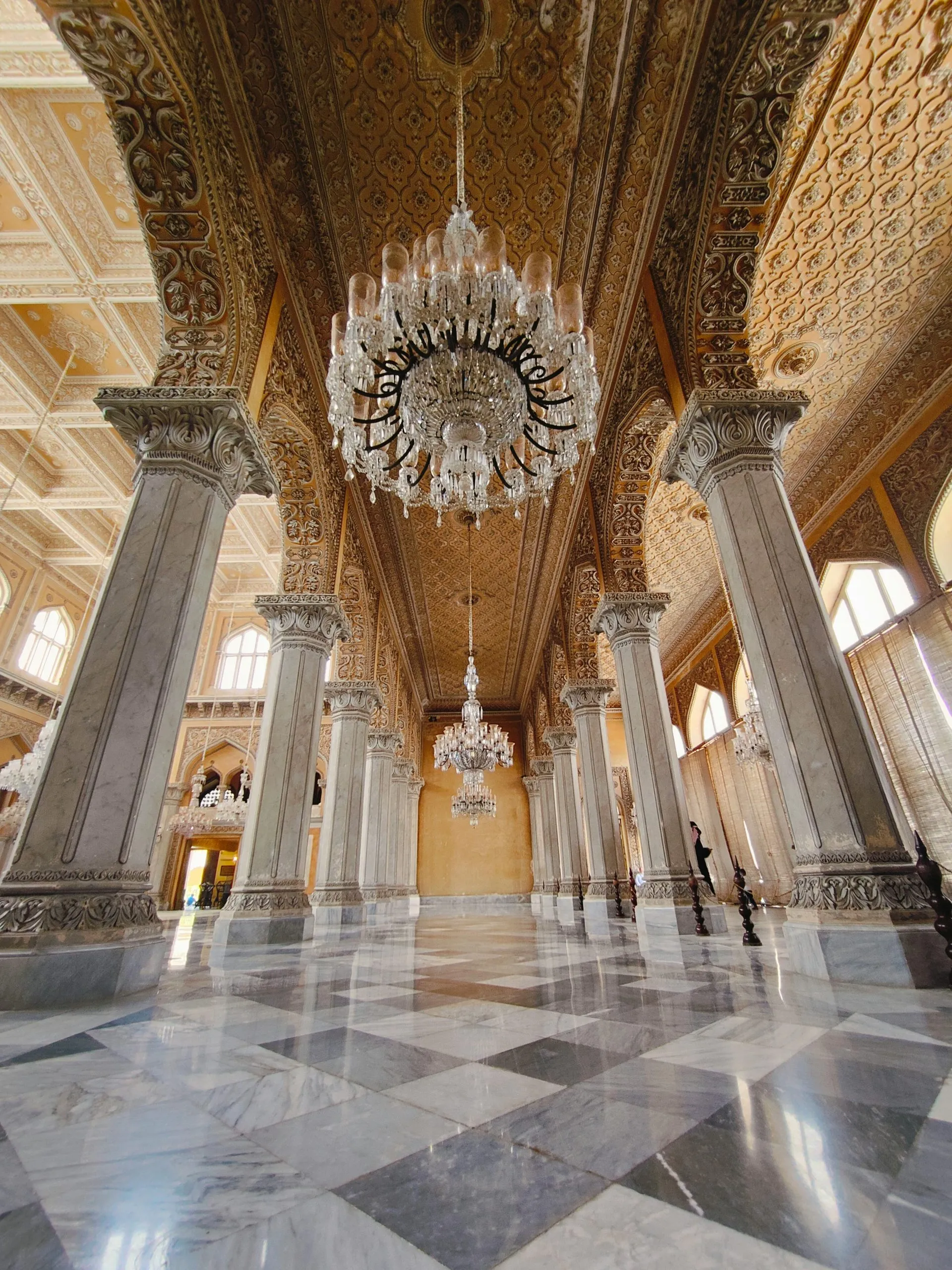 A grand hall with ornate columns, intricate golden ceilings, and elegant crystal chandeliers.