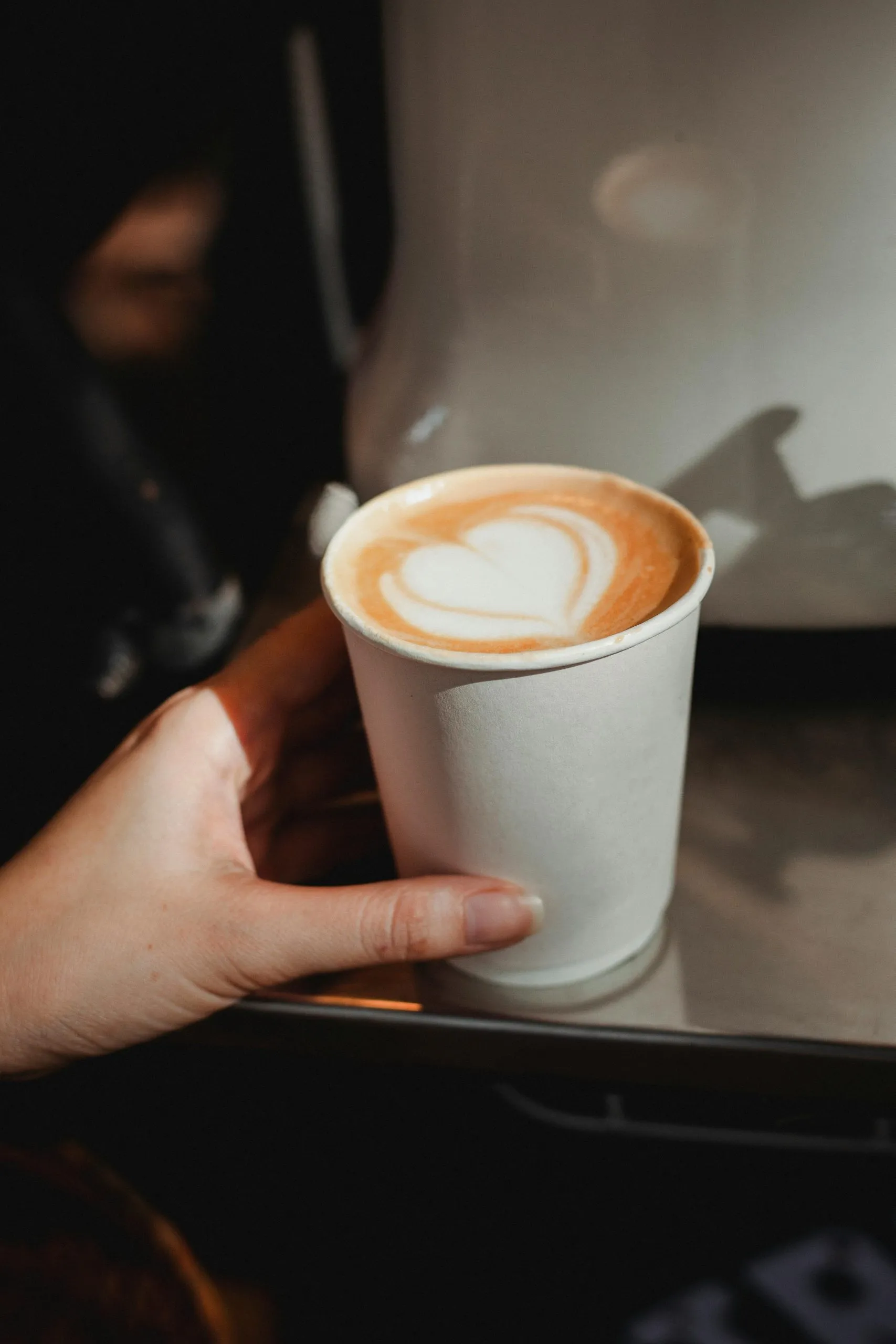 A hand holds a paper cup of latte with heart-shaped latte art, lit by soft, natural light.