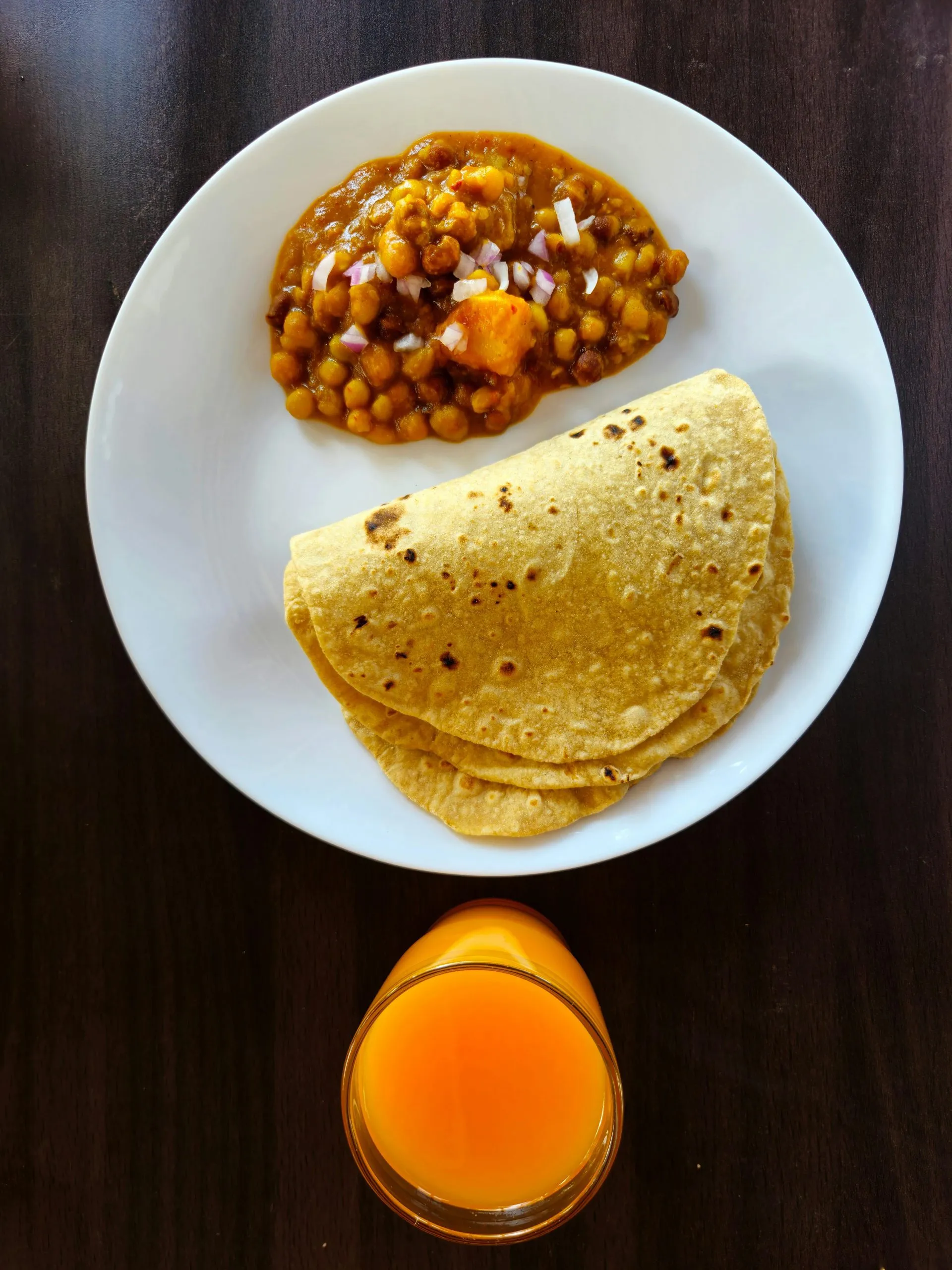 Plate with chickpea curry, chapati, and a glass of orange juice on a dark wooden table.