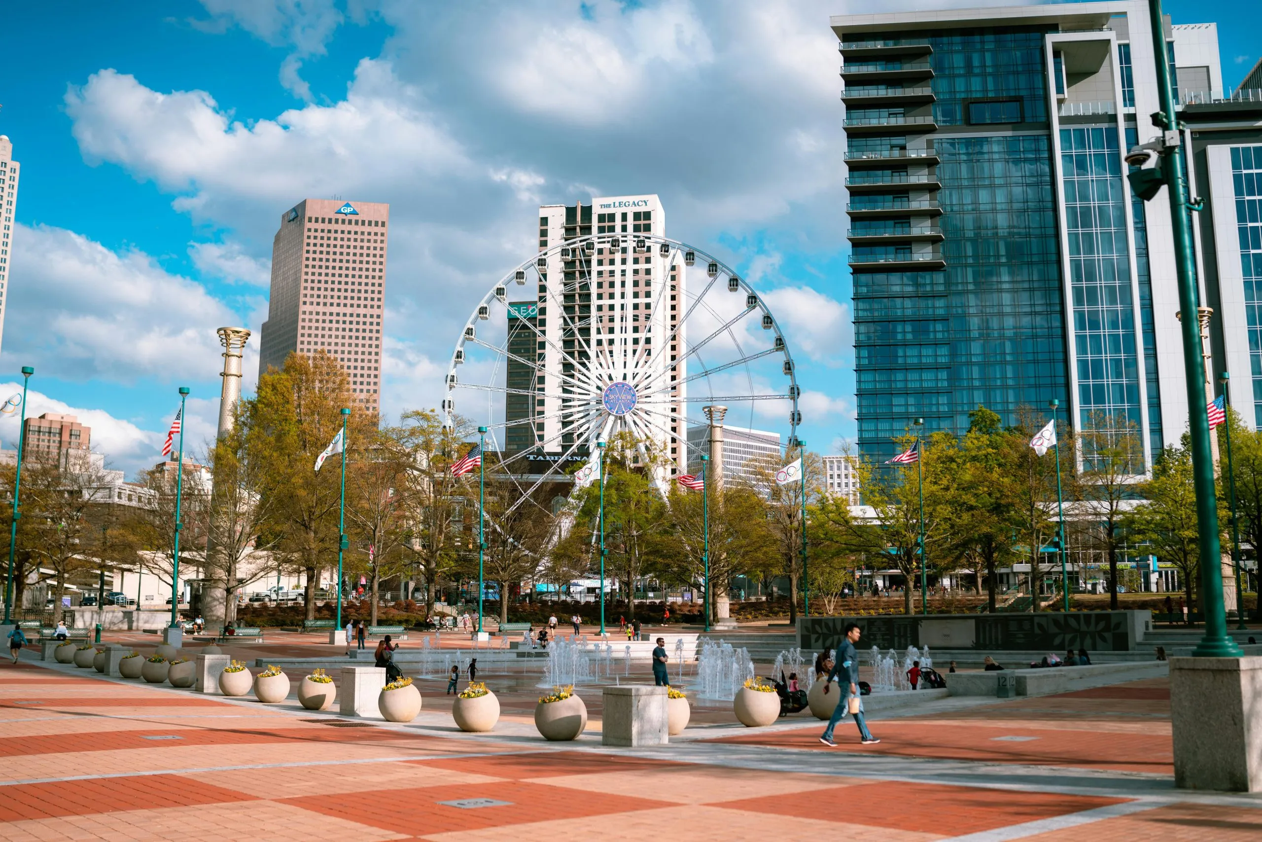 A cityscape featuring a Ferris wheel, modern buildings, and a public plaza with fountains.