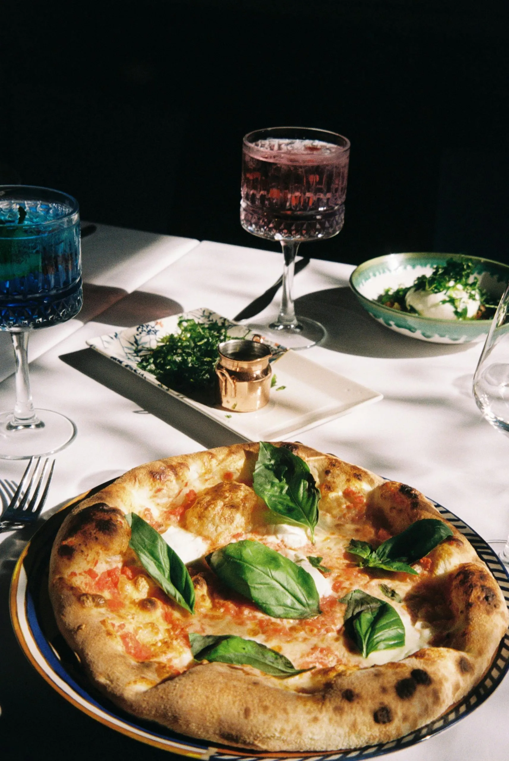 A Margherita pizza garnished with basil, paired with colorful drinks and side dishes on a table.