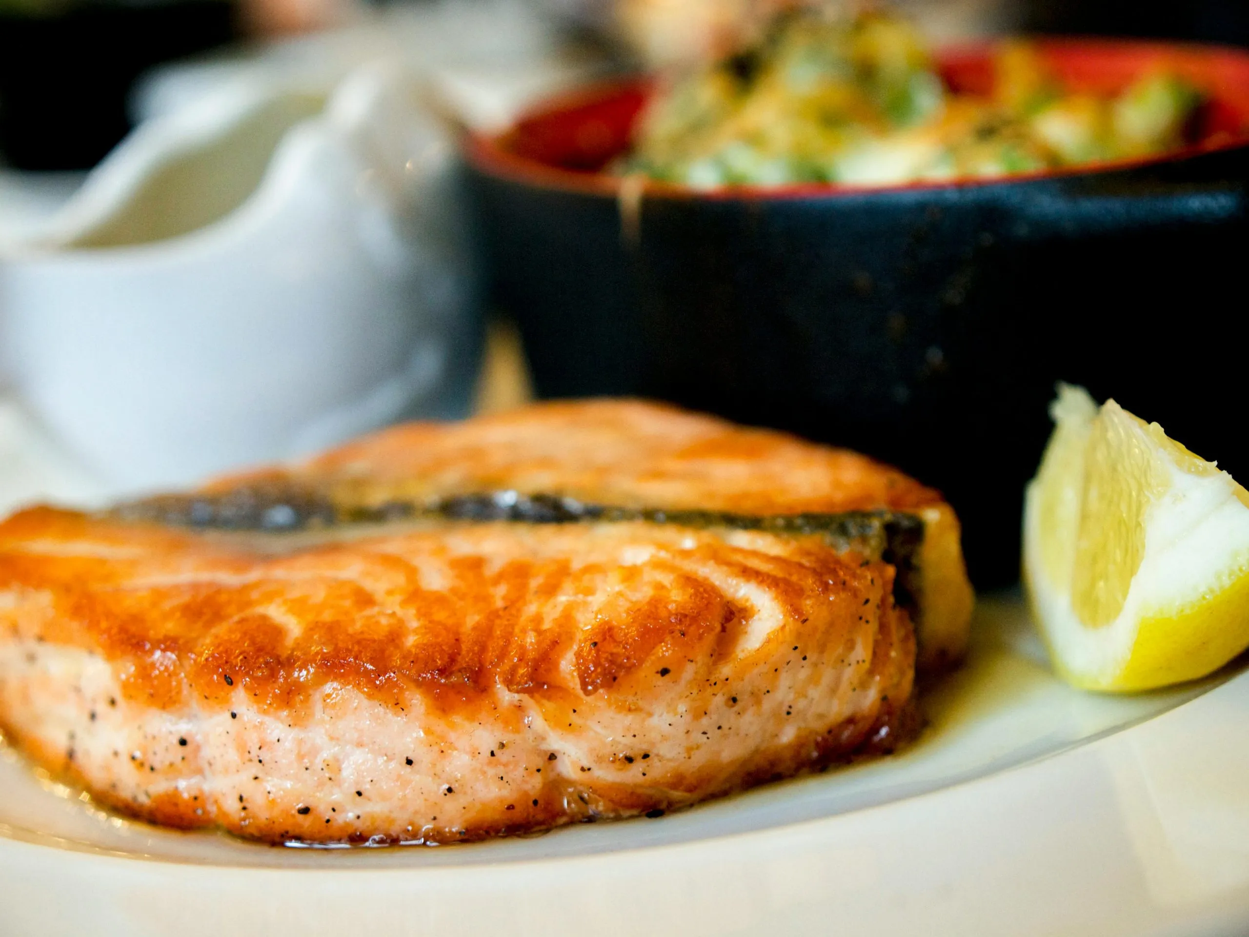 Seared salmon fillet served with a lemon wedge and a side dish in a black bowl.