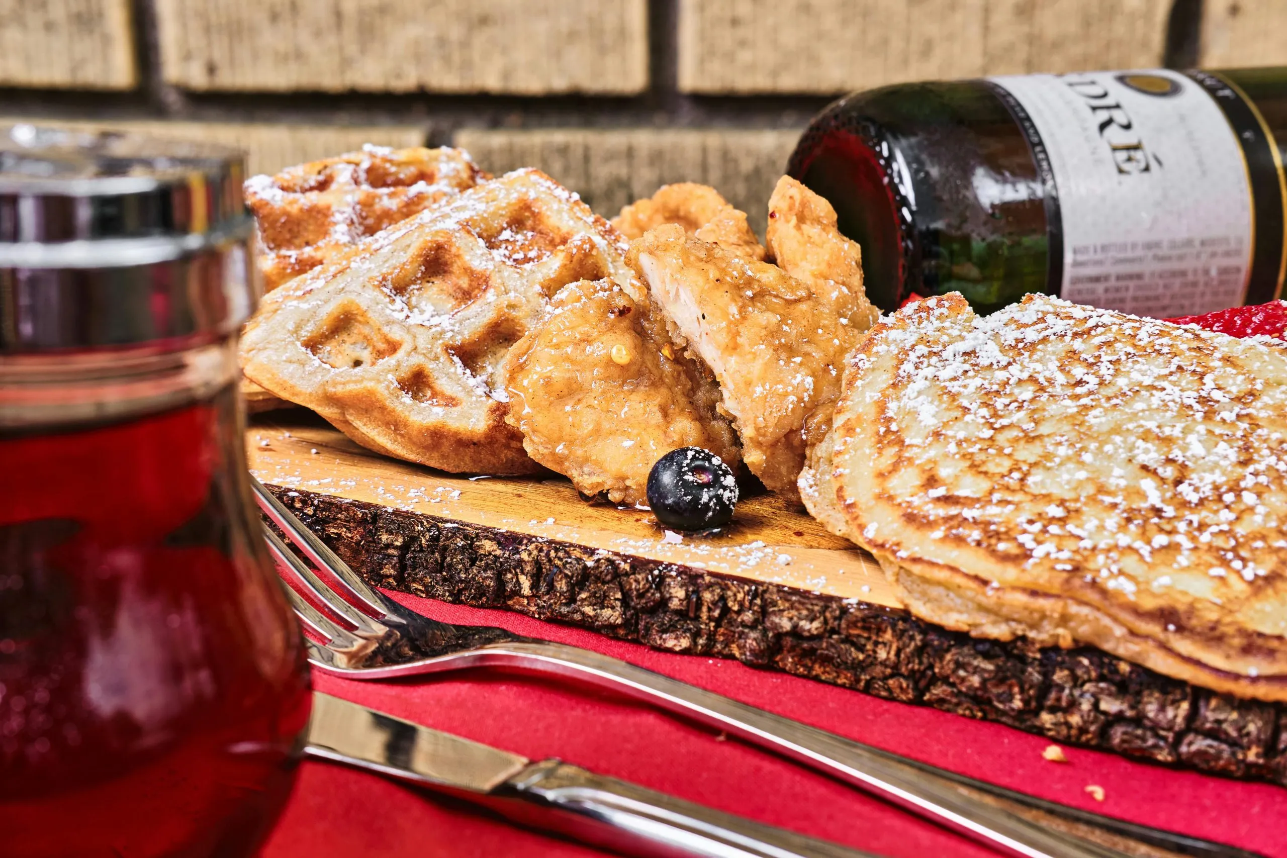 Waffles, pancakes, fried chicken, and syrup served on a rustic wooden platter with a wine bottle.