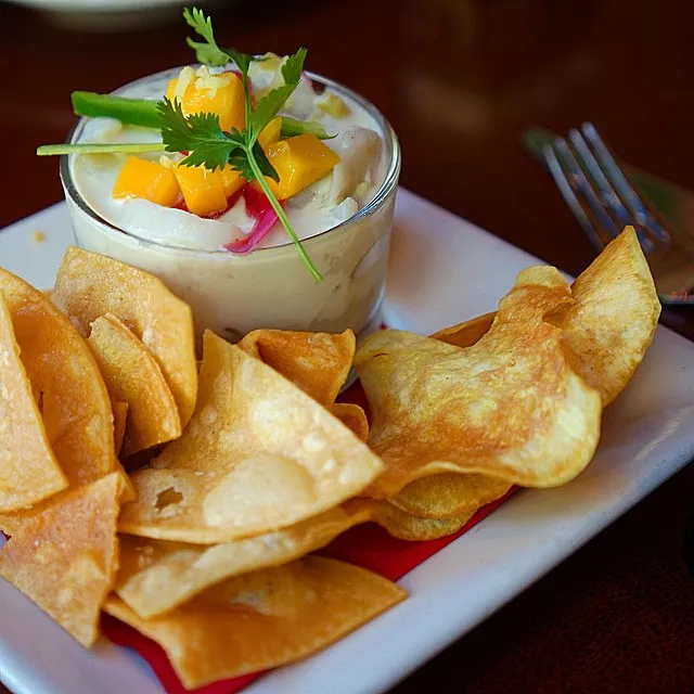 A creamy dip garnished with mango and herbs, served with crispy tortilla chips on a white plate.