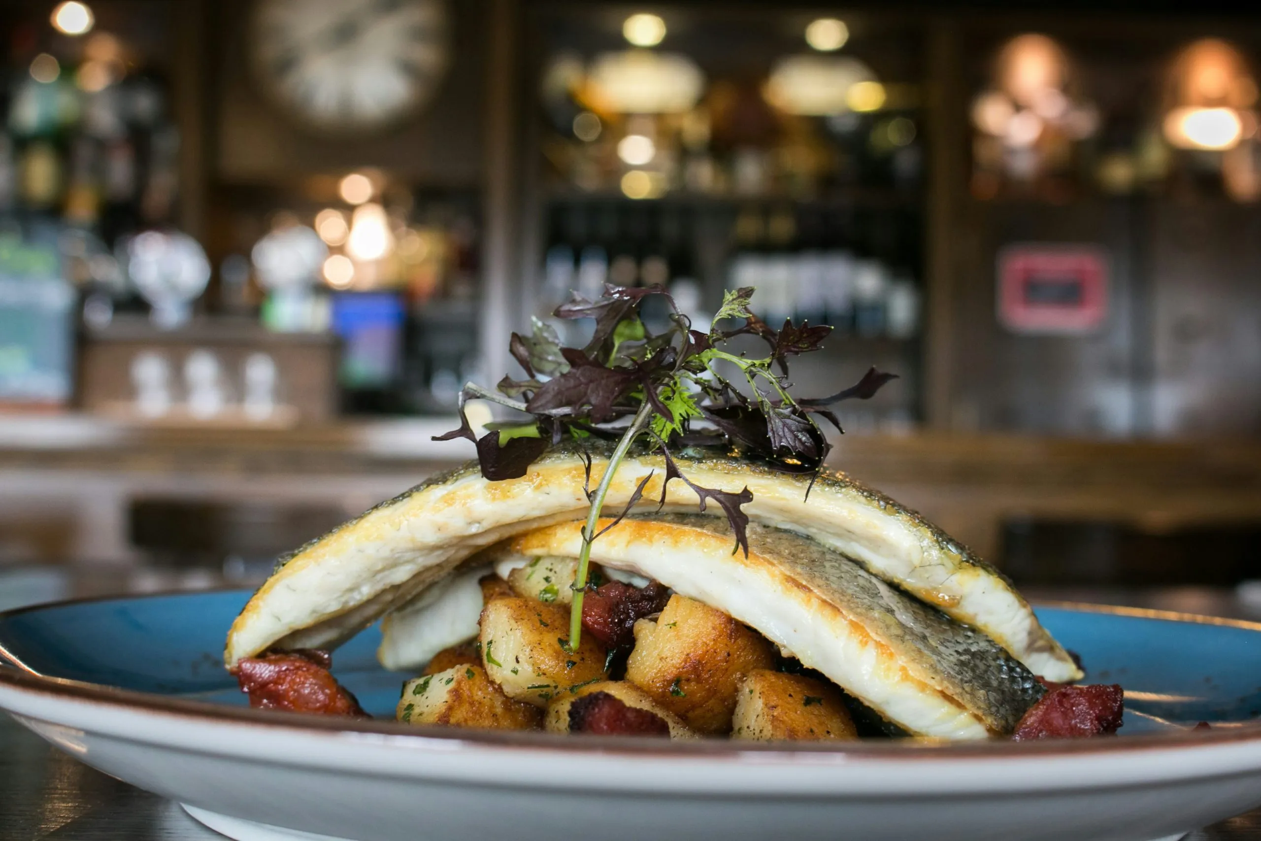 A plated dish featuring seared fish, roasted potatoes, herbs, and microgreens in a rustic setting.