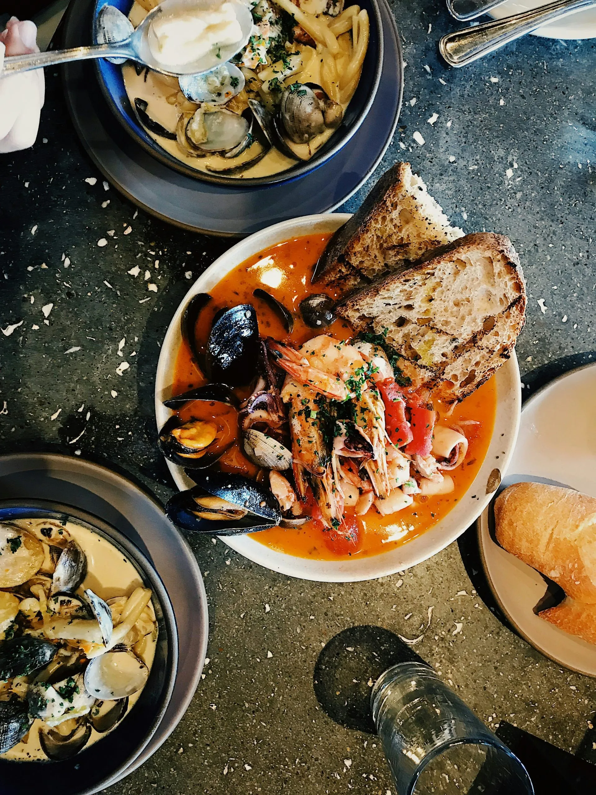 Seafood dishes with clams, mussels, shrimp, and bread served in bowls on a textured table surface.