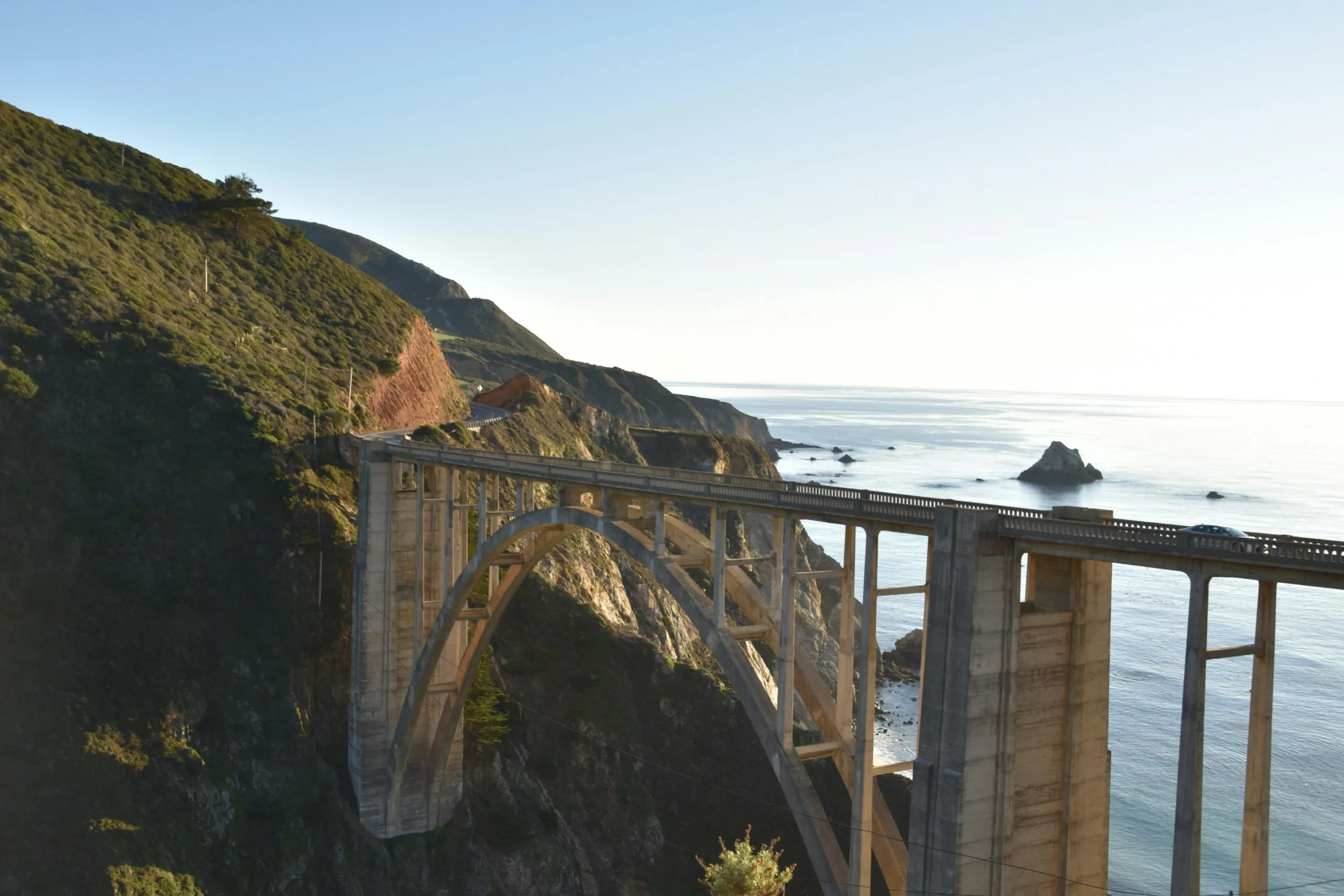 A coastal arch bridge spans a rugged cliffside, overlooking the serene ocean and rocky shoreline.