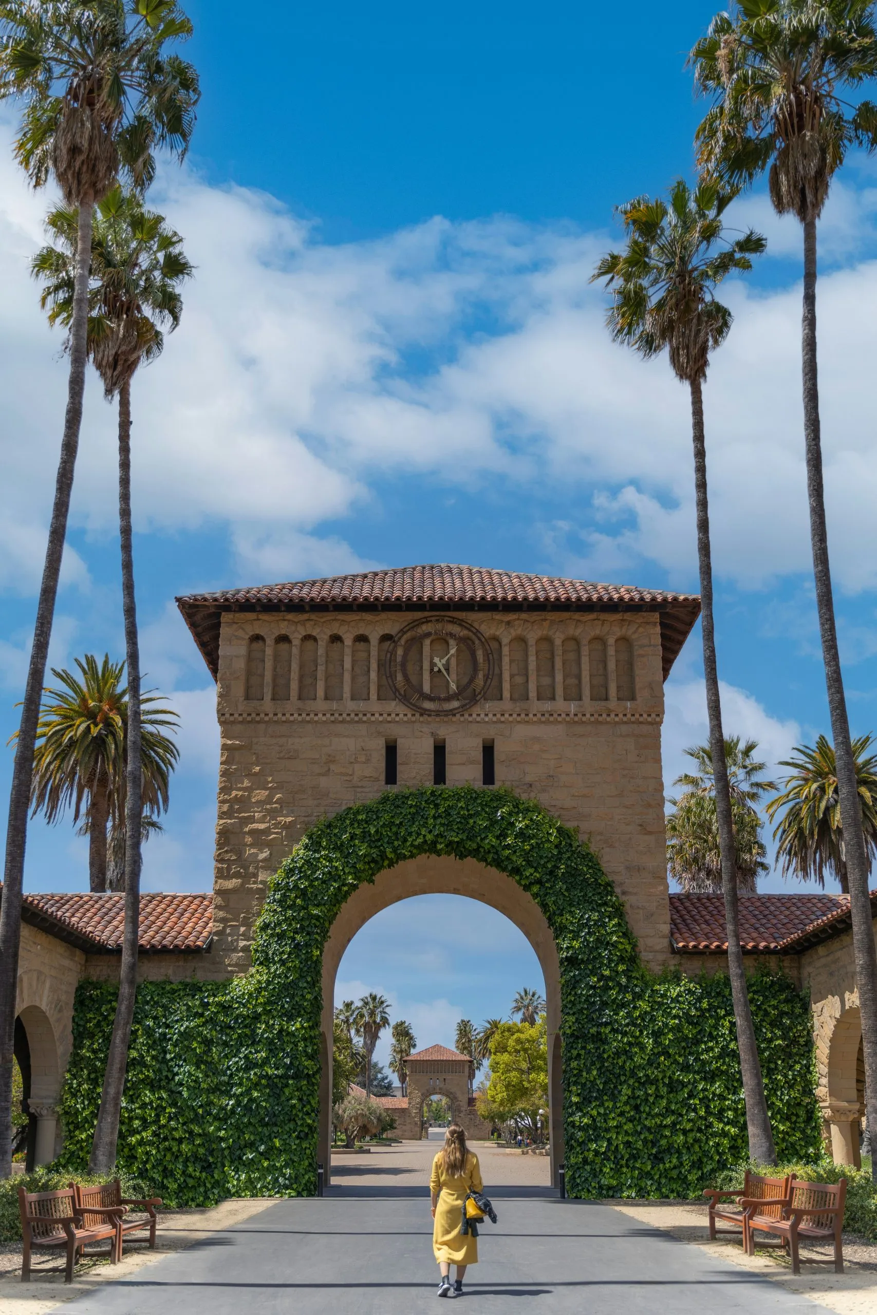 A woman in a yellow coat walks toward an ivy-covered archway surrounded by palm trees.