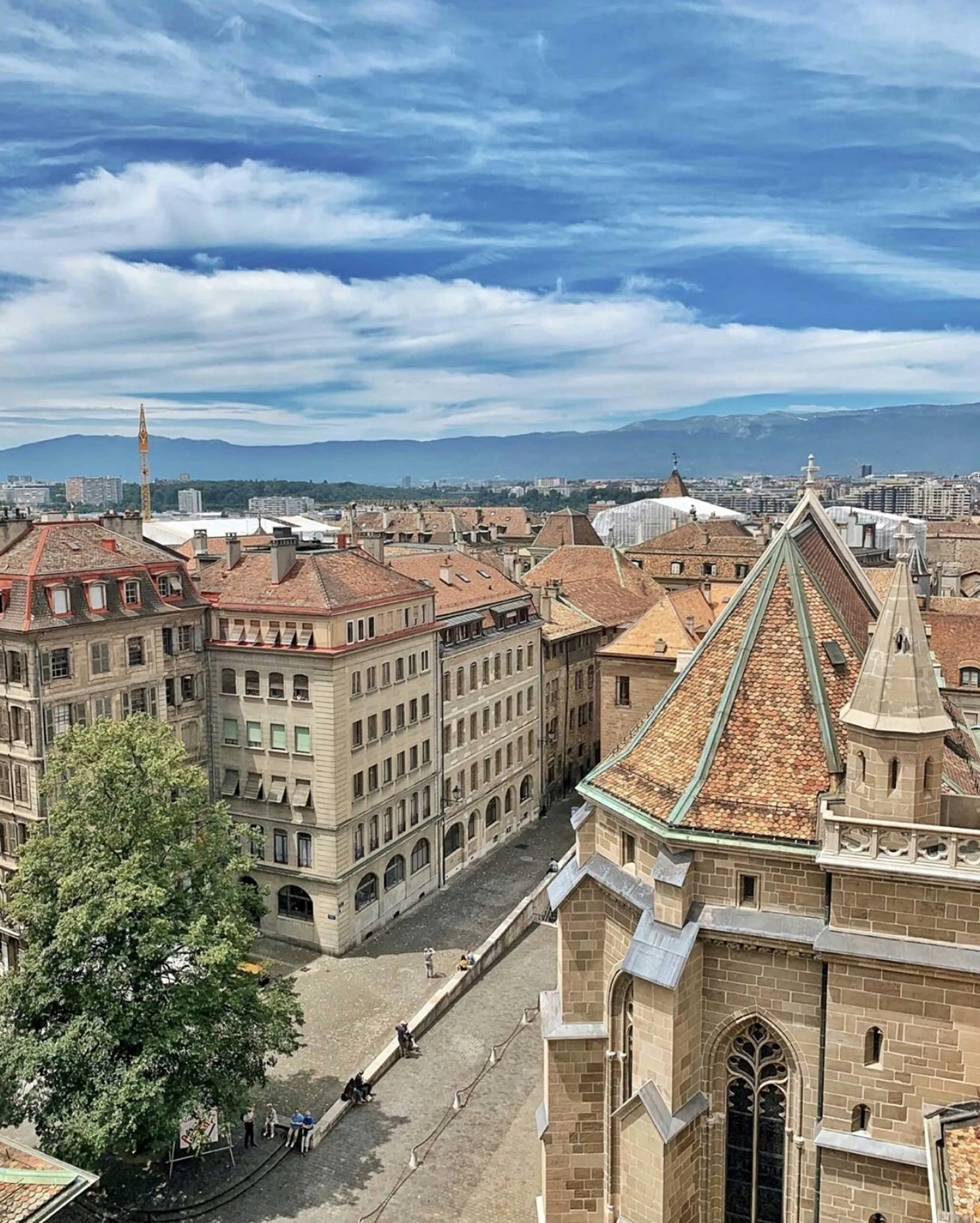 A historic European cityscape with tiled rooftops, Gothic architecture, and distant mountains.