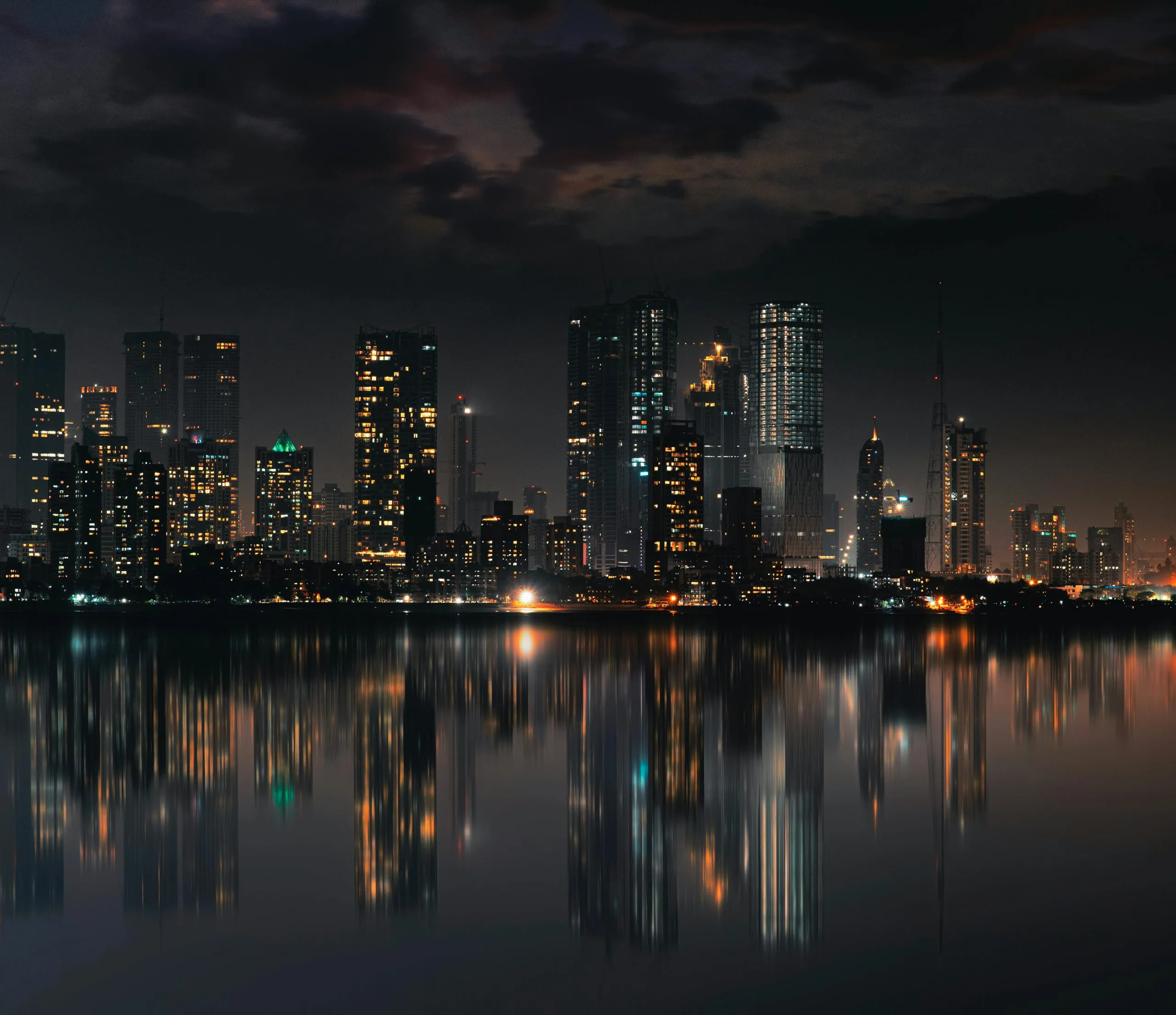 A city skyline at night reflects on calm water, illuminated by glowing lights and dark clouds above.