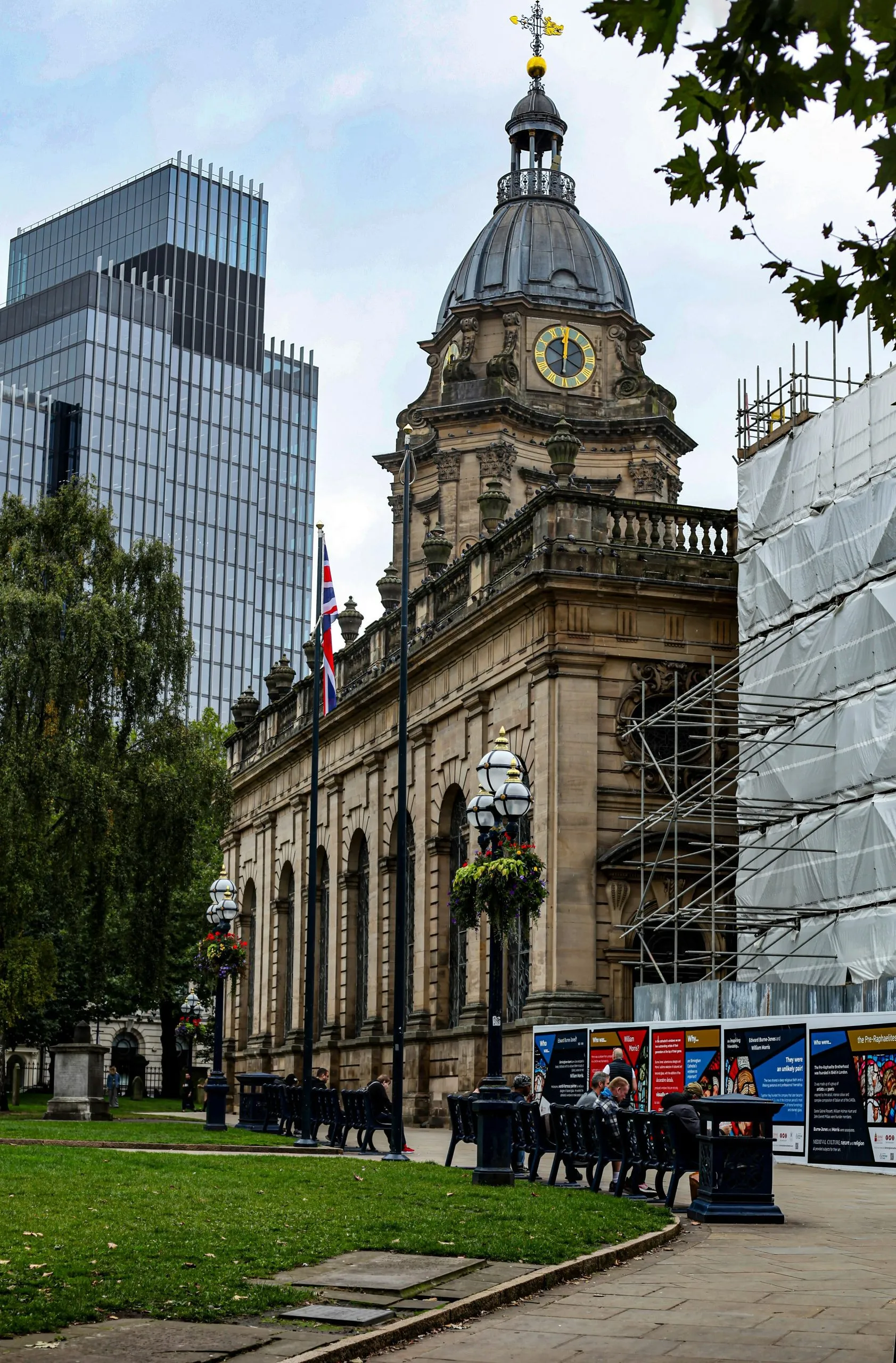 Historic building with a clock tower, modern skyscraper, and people seated in a green urban space.