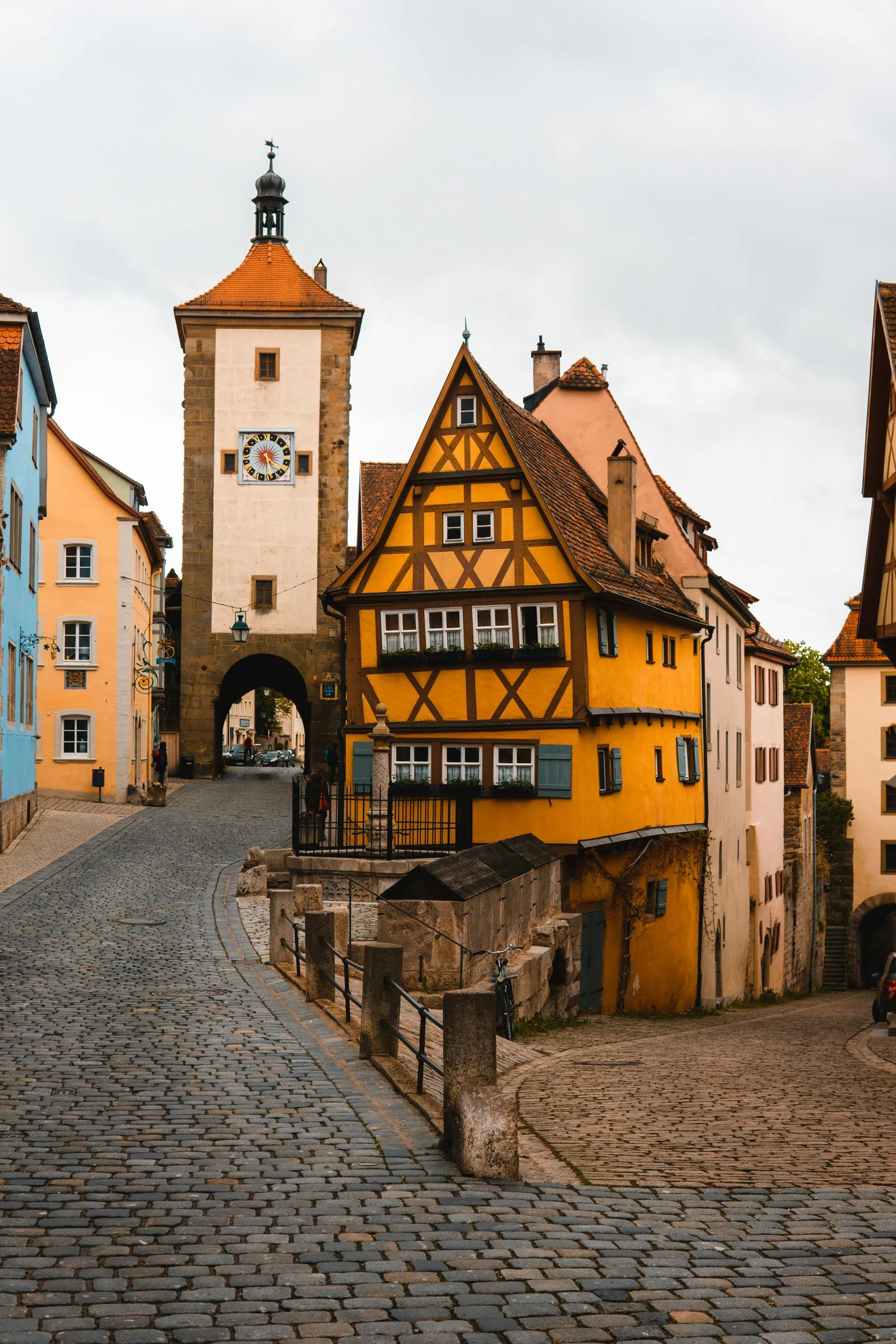 A cobblestone street leads to a medieval clock tower, flanked by colorful, half-timbered houses.