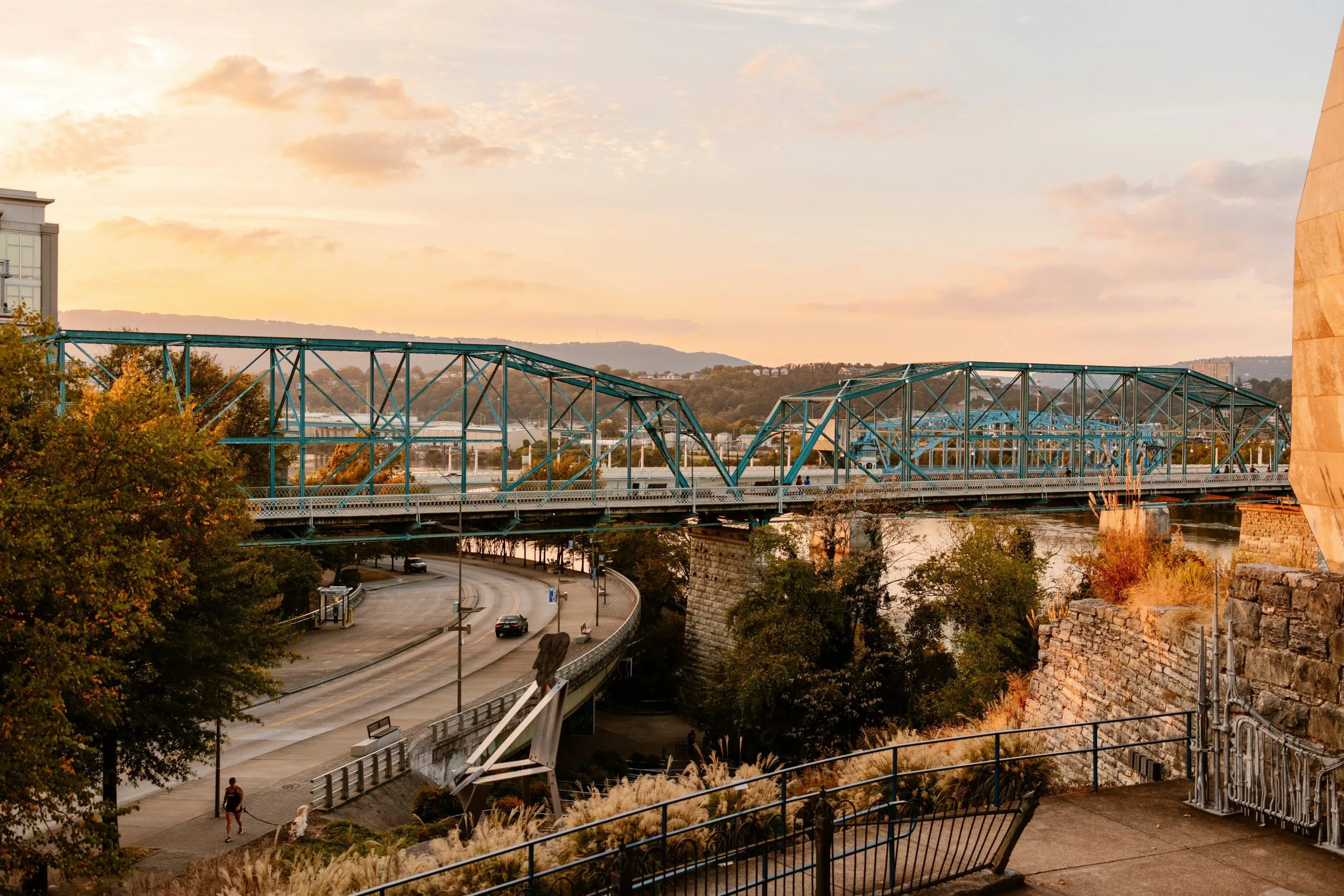 A blue truss bridge spans a river at sunset, surrounded by trees and urban pathways.