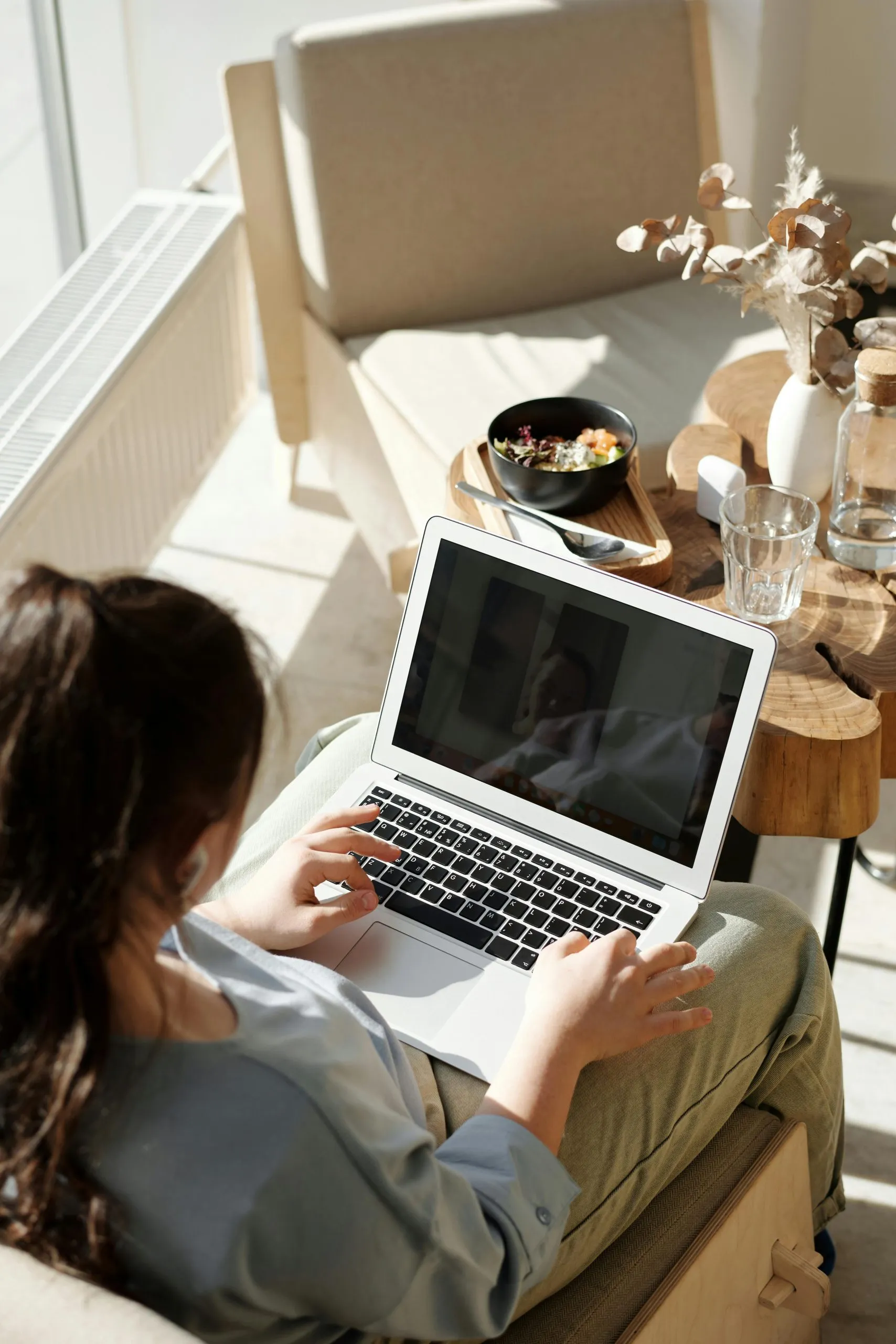 A person works on a laptop while seated near a table with food, water, and decor in natural light.