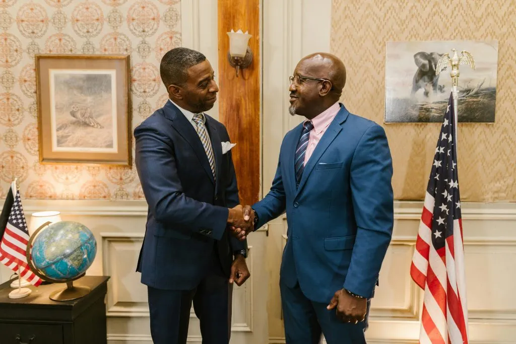 Two men in suits shake hands in a formal setting with U.S. flags and a globe in the background.