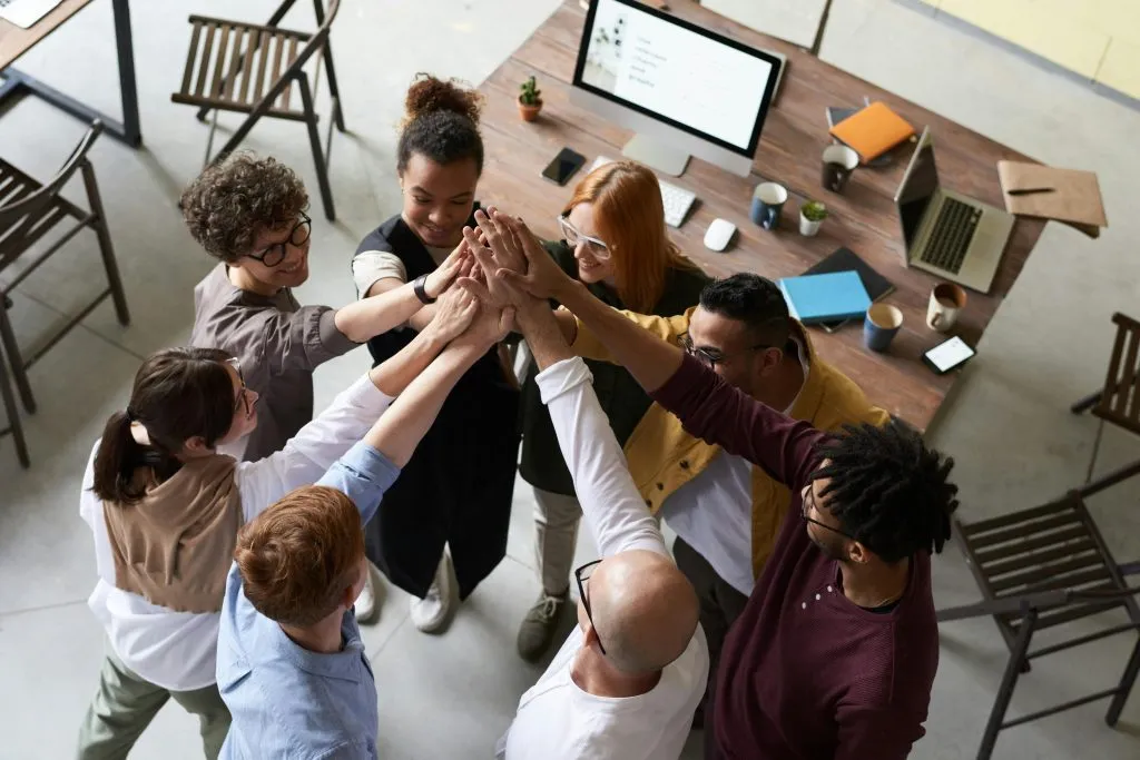 A diverse group of colleagues joins hands in a teamwork gesture around a workspace with laptops and notes.