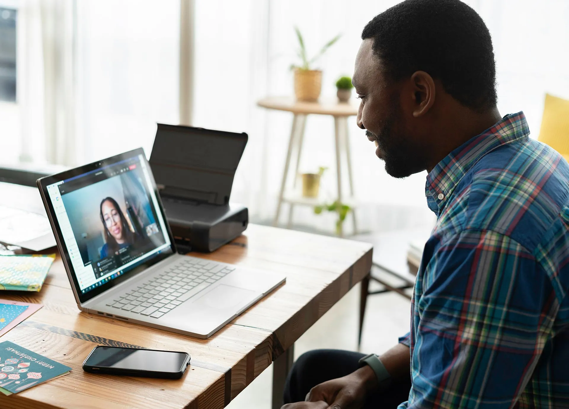 A person in a plaid shirt participates in a video call on a laptop at a wooden desk.