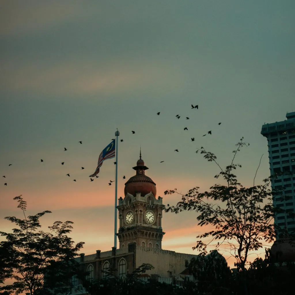 A clock tower with a Malaysian flag at sunset, surrounded by trees and flying birds.