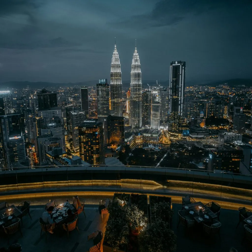 A nighttime cityscape featuring the illuminated Petronas Towers and a rooftop dining area.