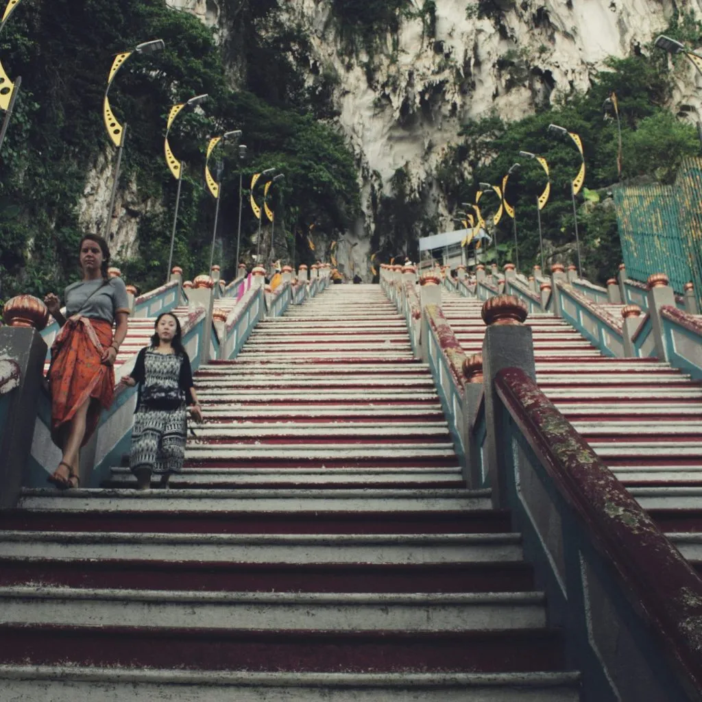 Steep stairs lead to a limestone cave, flanked by greenery and decorative yellow flags.