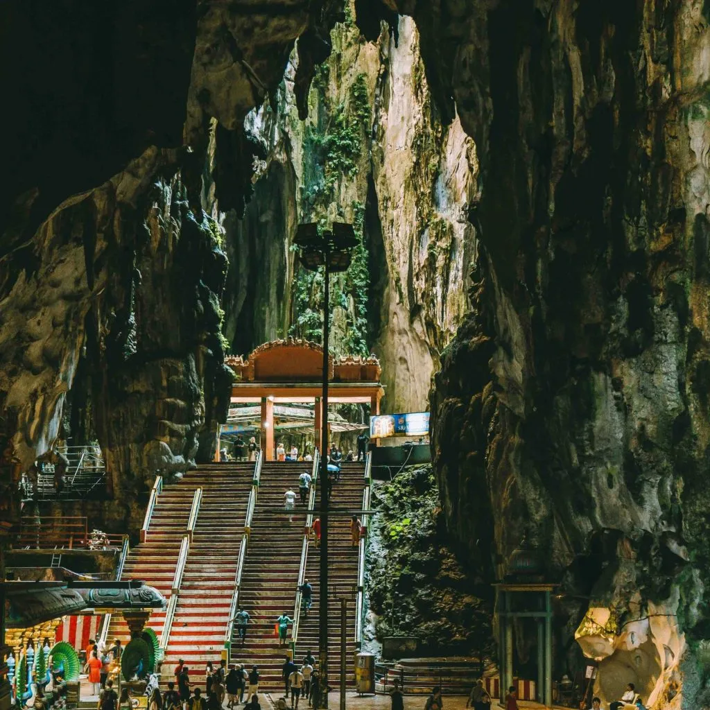 A grand staircase leads to a temple inside a massive, natural cave with lush greenery and rock formations.