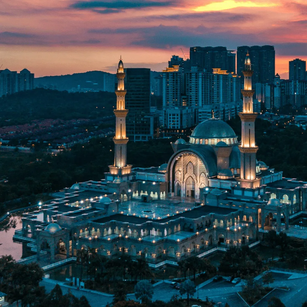 A grand mosque illuminated at sunset, surrounded by urban buildings and lush greenery.