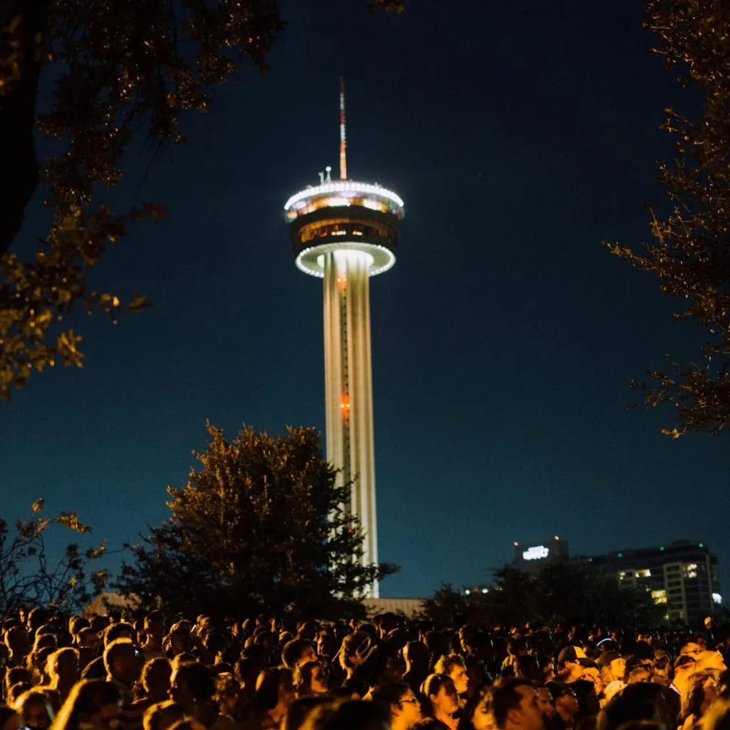 A crowd gathers at night near the illuminated Tower of the Americas, surrounded by trees and city lights.