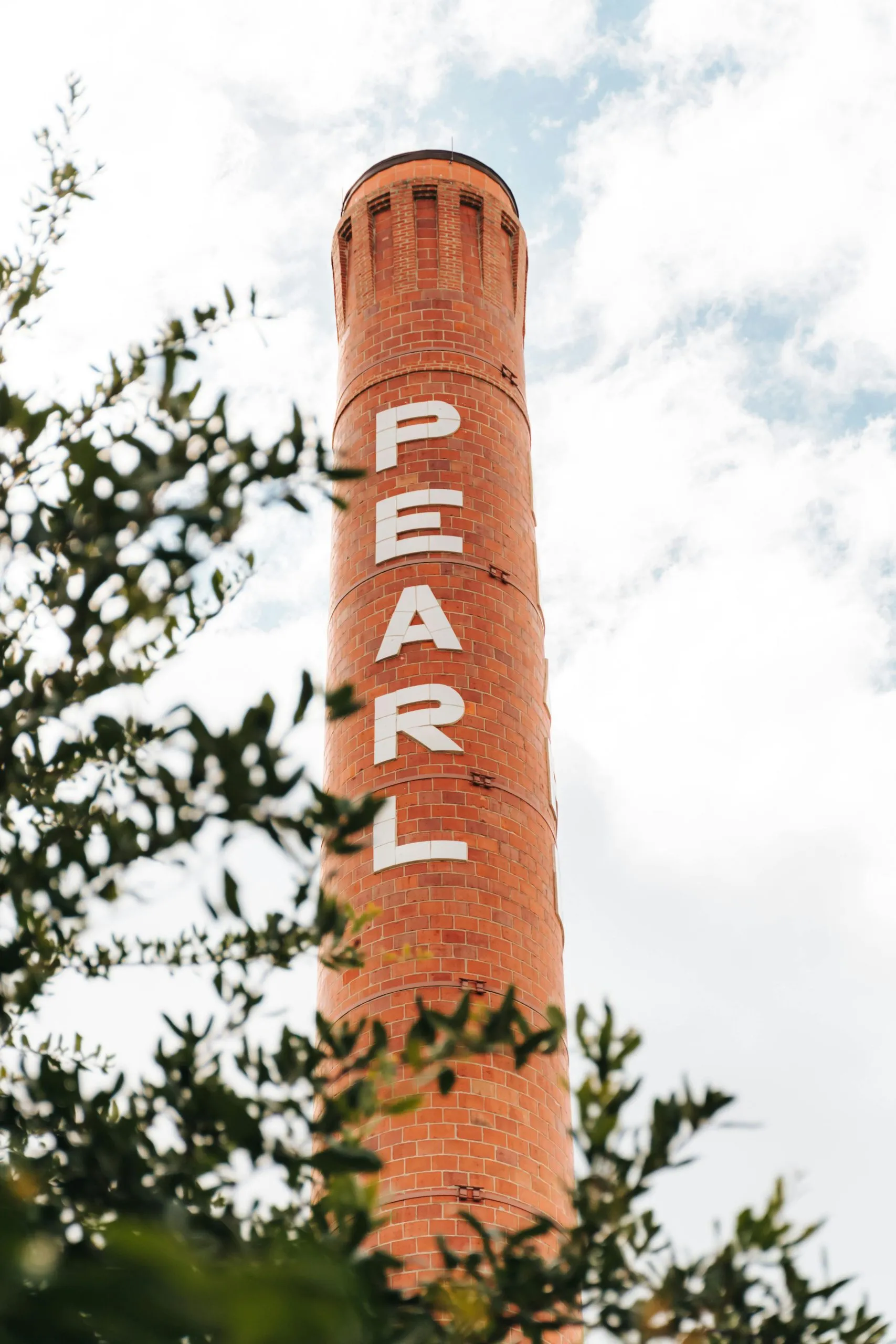 A tall red brick smokestack labeled "PEARL" rises against a cloudy sky, framed by leafy branches.