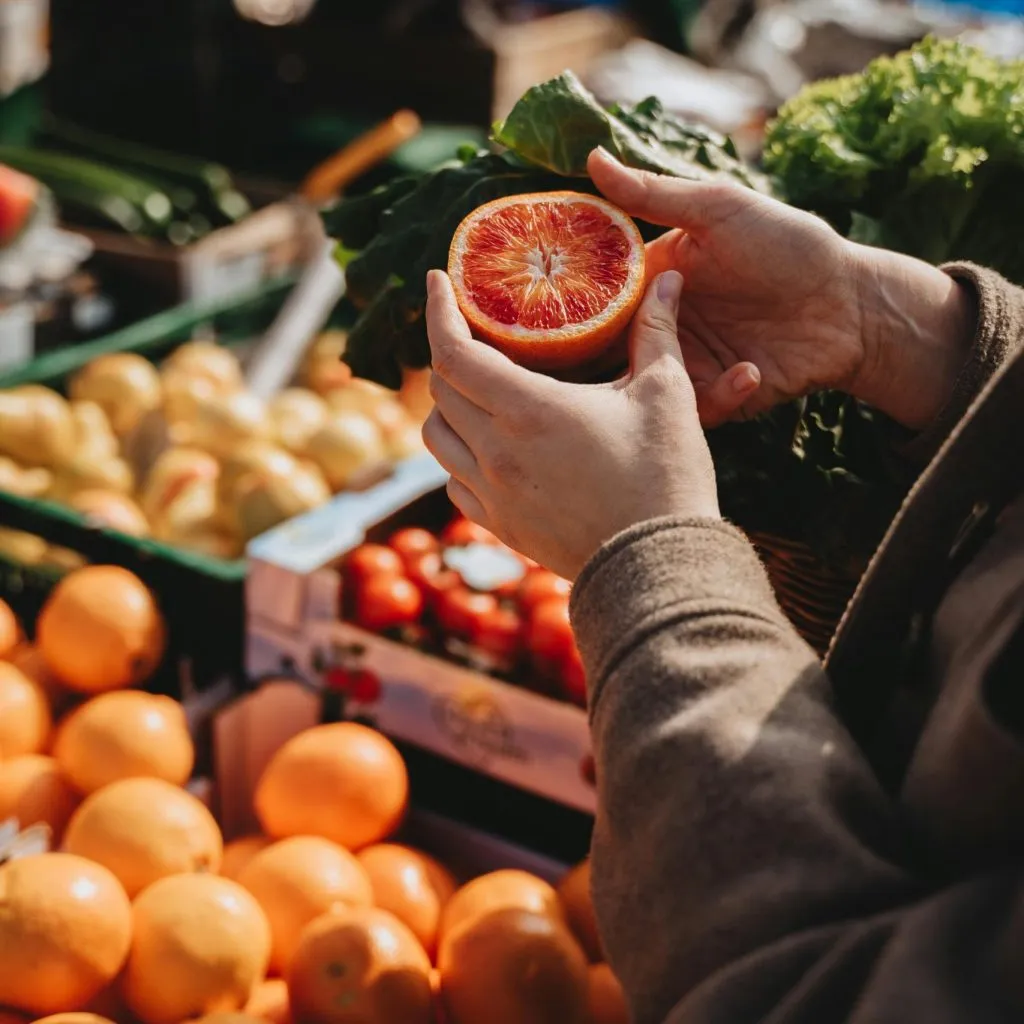 Hands holding a halved blood orange at a market stall with fresh produce in the background.