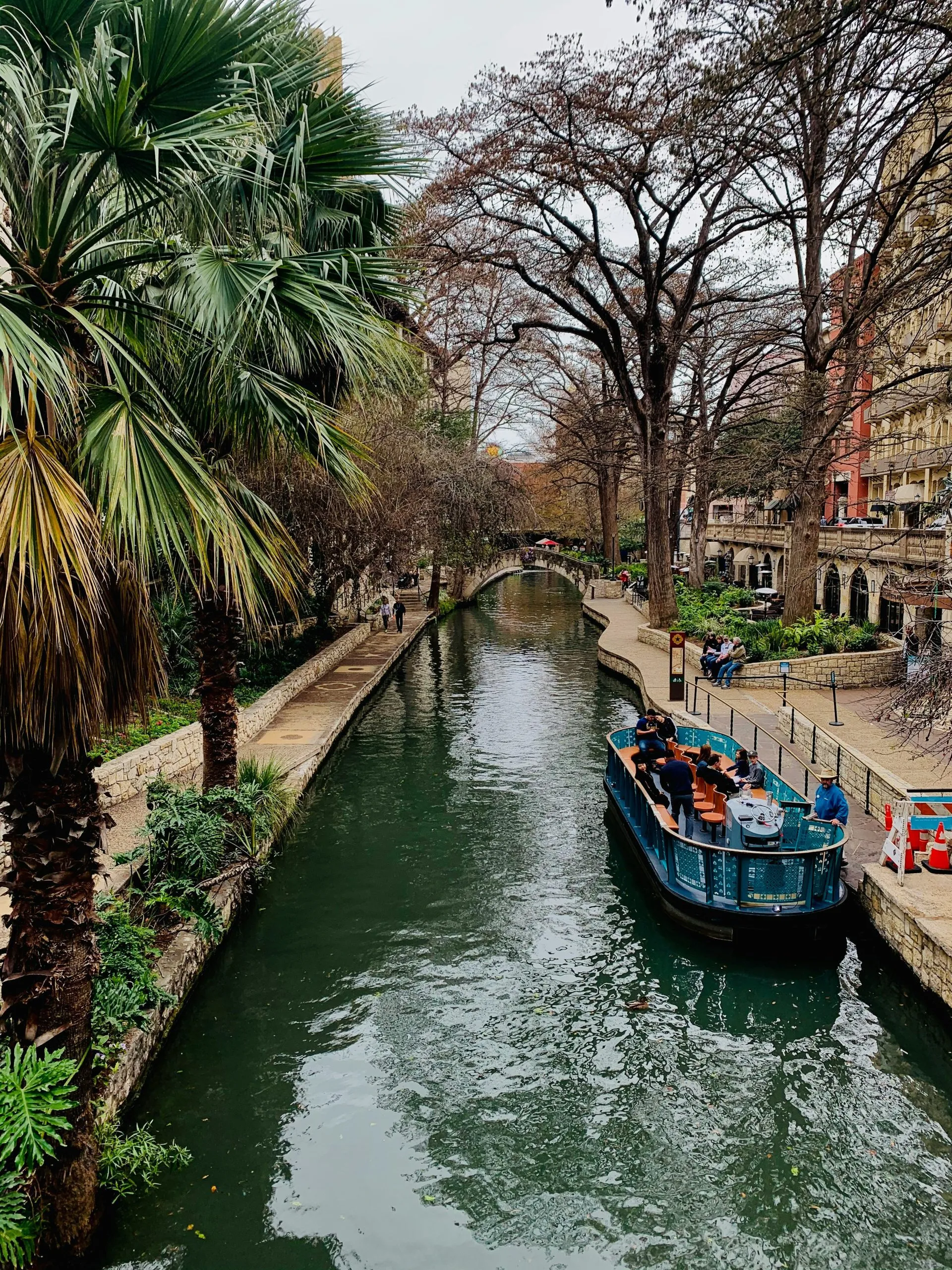 A scenic canal lined with trees, a boat carrying passengers, and a walkway with pedestrians.