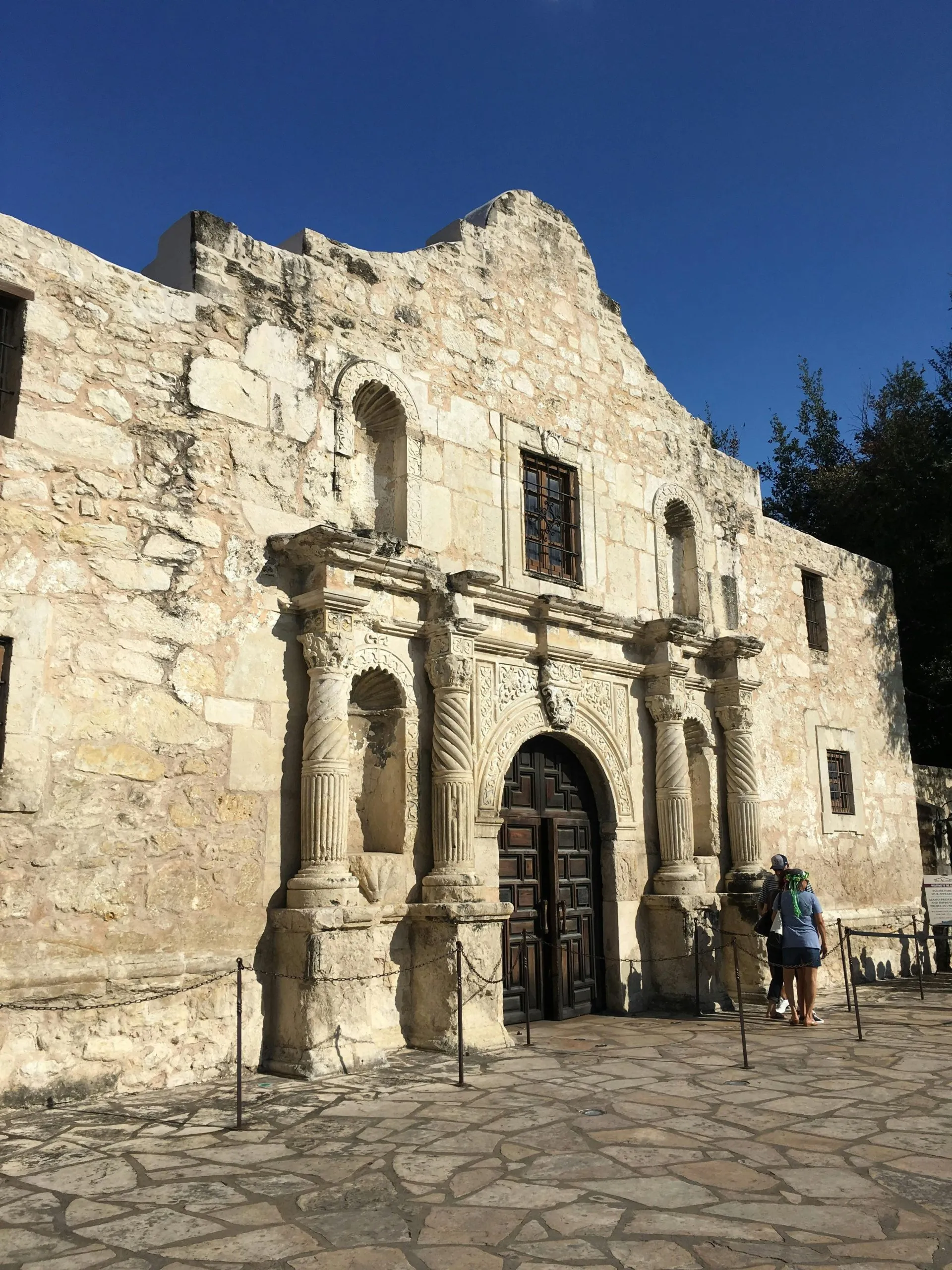 The Alamo's historic stone facade under a clear blue sky, with visitors standing near the entrance.