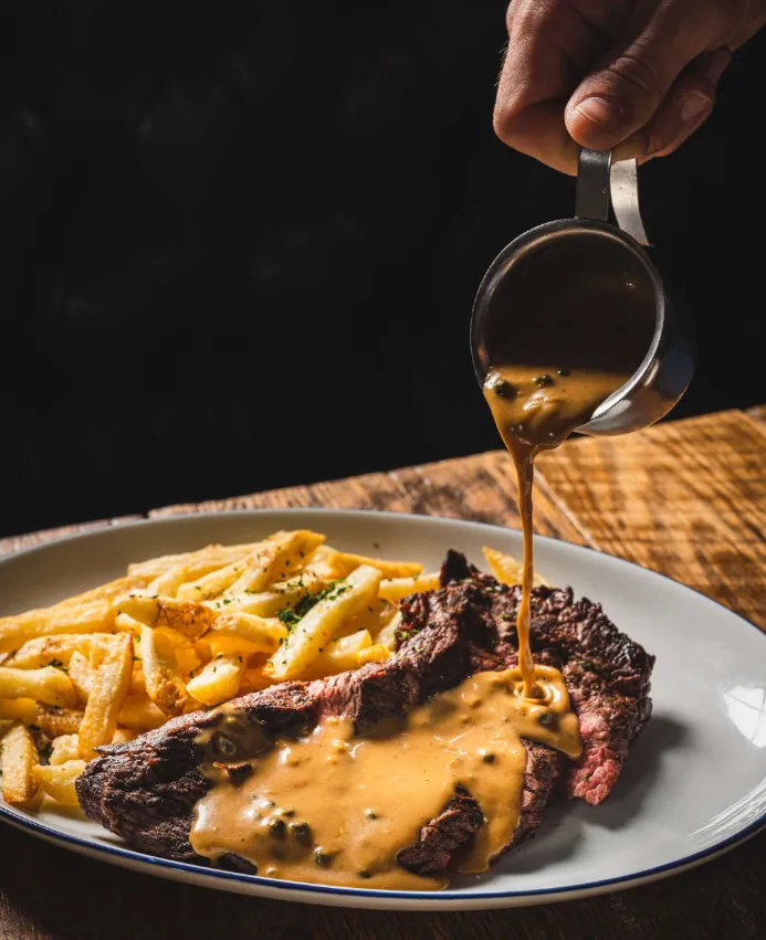 A steak topped with creamy sauce is served alongside seasoned fries on a white plate.