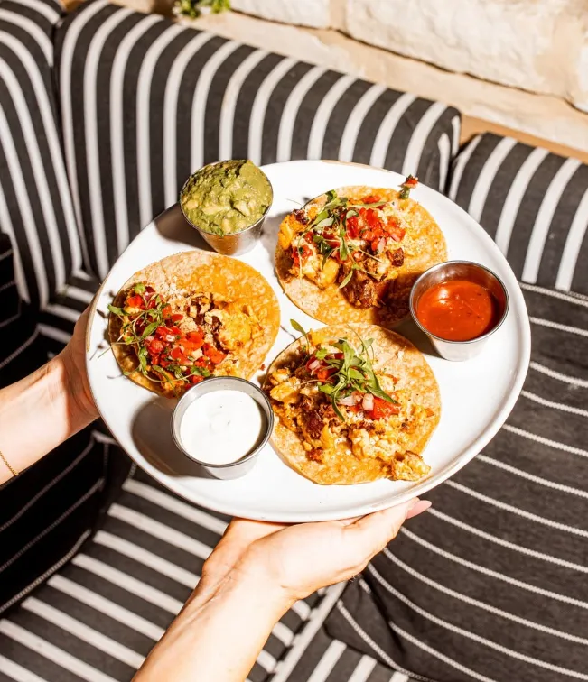 A plate of tacos with guacamole, salsa, and sour cream, served on a striped seating area.